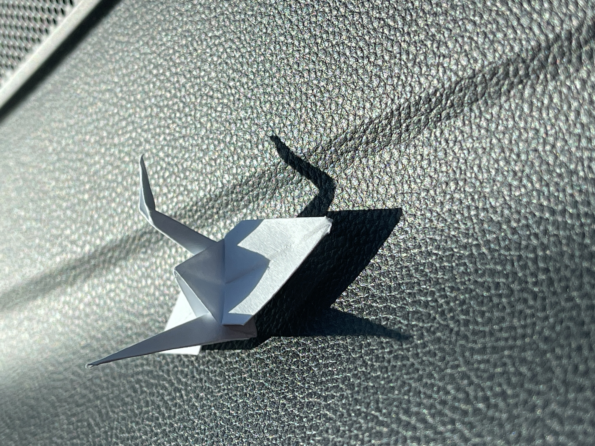 A white origami paper model, resembling a stylized paper airplane or bird with sharp folds and upward-pointing elements, is the central focus. It rests on a dark gray, heavily textured surface, similar to pebbled leather or a durable plastic interior. The texture creates numerous small, glinting highlights under the strong light.
Direct sunlight from the upper left casts distinct, elongated shadows of the origami piece onto the textured background, revealing the intricate angles and depth of the paper folds. In the upper left portion of the image, a narrow, perforated grille or vent, possibly part of a car's dashboard or air conditioning system, is partially visible.
The sharp shadows and bright highlights indicate a sunny day, likely taken during mid-morning or mid-afternoon. The setting suggests the interior of a vehicle. There is no activity or human presence, and no visible text. The photograph captures this still life in Yastrebovo, Bulgaria.