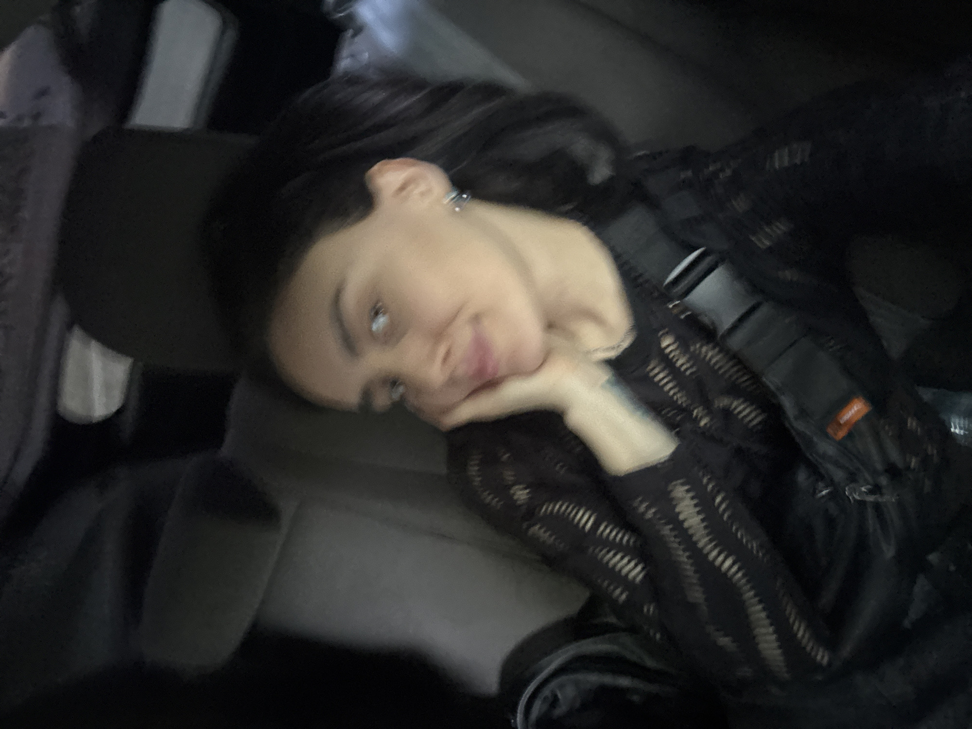 A woman with long dark hair and fair skin is resting her head on her hand, appearing to be in the back seat of a car. She is wearing a black long-sleeved shirt with a patterned mesh design, along with a black backpack or vest. The background is blurry, suggesting motion or a shallow depth of field. The location is unknown; there is no information in the image to suggest it is in Tbilisi, Georgia, or any other specific city.