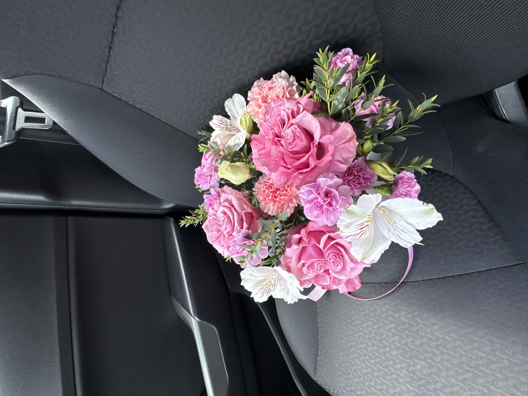 A bouquet of pink and white flowers rests on the black fabric seat of a car, likely a vehicle parked in the city of Izhevsk, Russia. The bouquet is comprised of various types of flowers, including roses, carnations, and alstroemerias, with green foliage interspersed throughout. A thin pink ribbon is visible, loosely wrapped around a portion of the bouquet. The car's interior, specifically the door panel and seatbelt buckle, are visible in the background. The lighting suggests it is daytime.