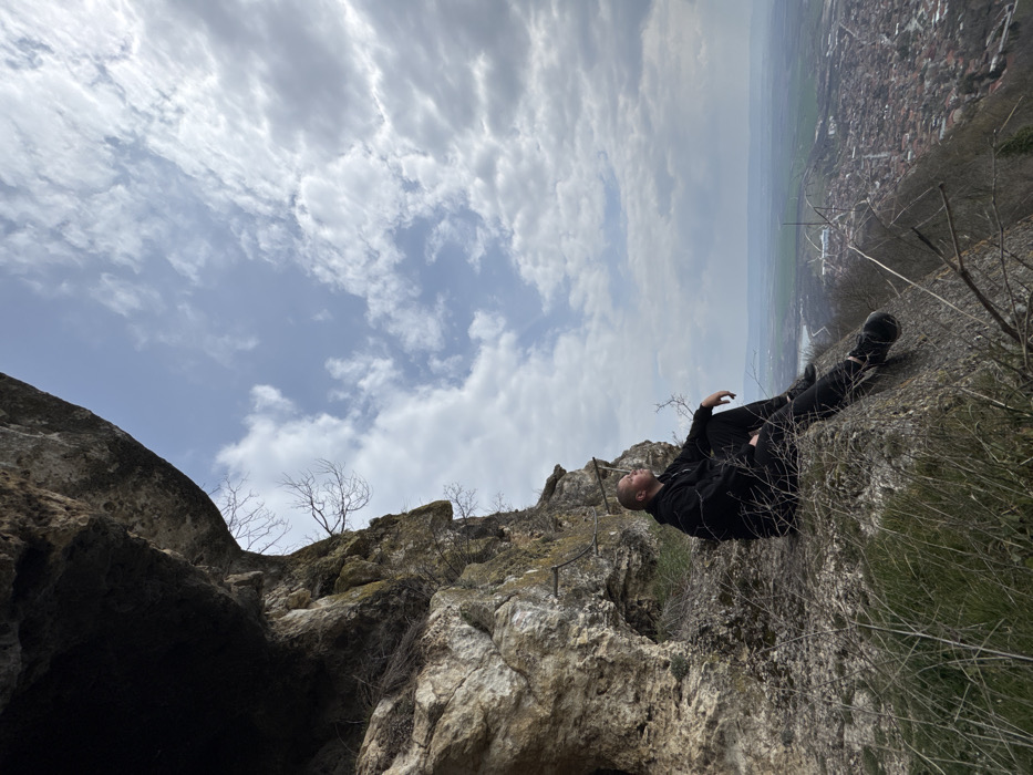 A male individual is seated on a weathered rock ledge beneath a substantial rock overhang on the Shumen Plateau, overlooking the city of Shumen, Bulgaria. The subject is dressed entirely in black, consisting of a hooded top, trousers, and athletic shoes. He is positioned facing right, with his legs extended forward and his head tilted upwards and to the right, observing the sky and distant horizon. His left hand rests on his left thigh. The immediate surrounding environment includes the rugged, light-colored rock face, sparse dry grasses, and bare tree branches. Below and extending into the distance, a panoramic view of Shumen city is visible, characterized by dense urban structures and cultivated fields, leading to a hazy horizon. The sky above is partly cloudy, displaying patches of blue sky interspersed with white and grey clouds.