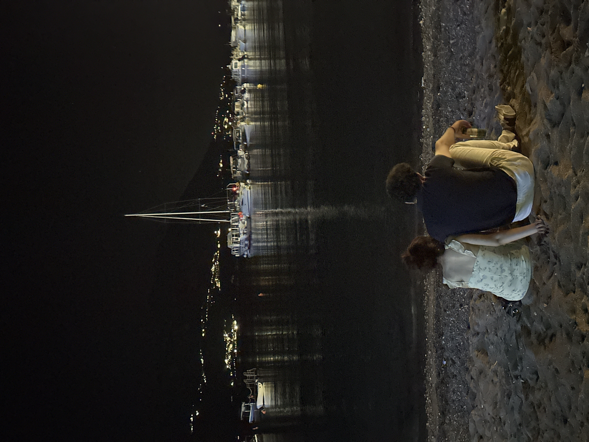 A man and a woman are sitting on a beach at night, facing away from the camera. They are looking out at a bay filled with boats, with a town visible on the hills in the distance. The man is wearing a dark t-shirt and light-colored pants, and the woman is wearing a floral dress. They appear to be a couple, and they are sitting close together. The water is calm and reflects the lights of the town. The beach is dark and sandy, with some pebbles visible. The scene is peaceful and romantic.