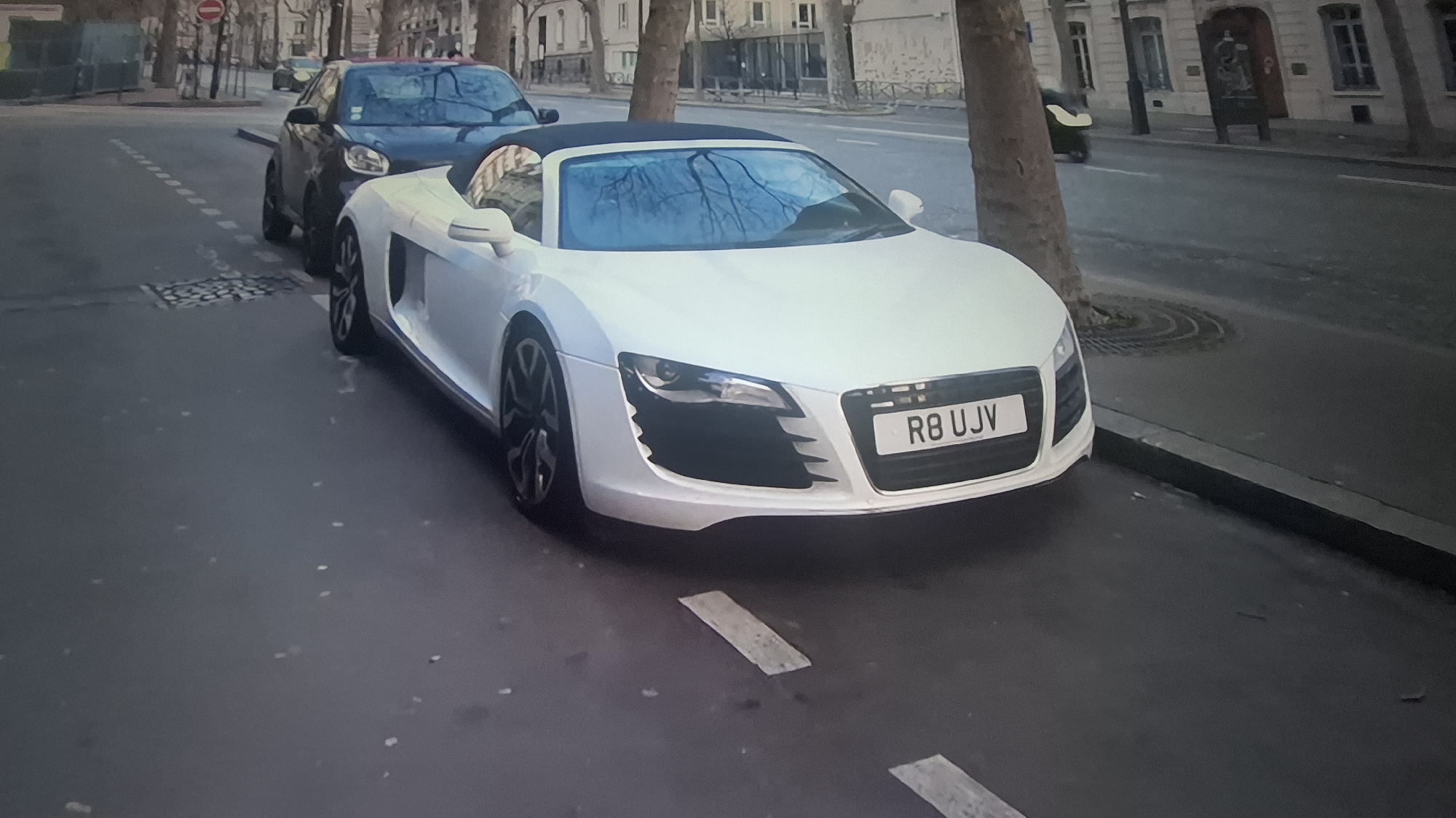 A white Audi R8 Spyder with license plate "R8 UJV" is parked on a city street. Another dark-colored compact car is parked behind it. There are trees lining the street, and buildings are visible in the background. The location appears to be urban, possibly a European city. A no-entry road sign can be seen in the background.