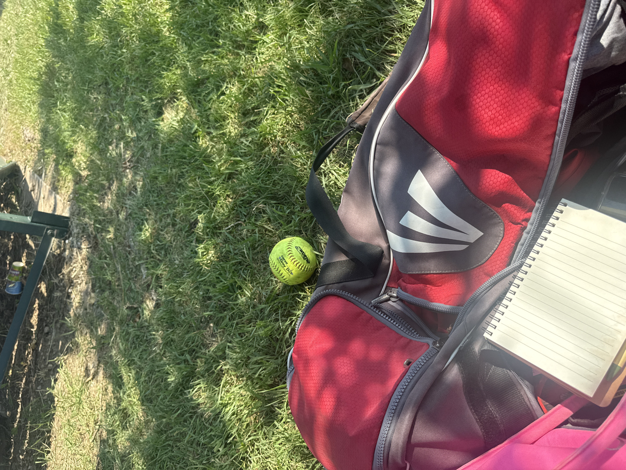 An eye-level, close-up shot shows a red and gray softball bag with an open zipper, lying on green grass. A yellow softball rests on the grass to the left of the bag. Inside the bag, a spiral-bound notebook is visible. The background shows dappled sunlight filtering through leaves onto the grass and the edge of a dark structure. The scene appears to be outdoors in a park or backyard setting.