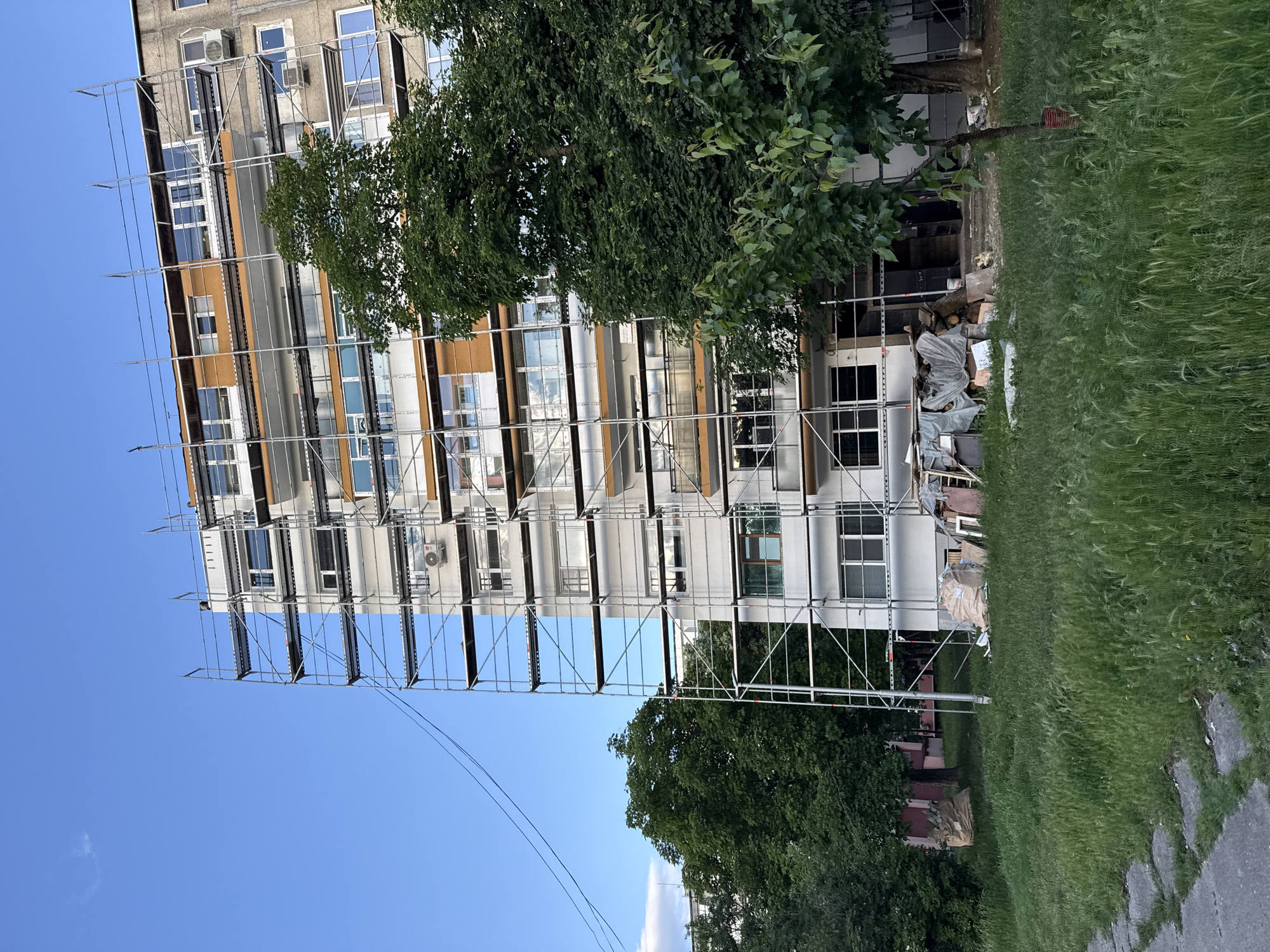 The image shows a tall, multi-story building under renovation, evidenced by the scaffolding erected along its façade. The building's exterior is primarily white, with some sections appearing tan or beige. Numerous windows are visible, some of which have air conditioning units installed.
The scaffolding is made of metal and extends nearly to the full height of the building.
Adjacent to the building, there's a grassy area with trees. Debris and construction materials are also present on the ground.
The sky is blue. The image was taken during daylight.
This is a building located in Lovech, Bulgaria.