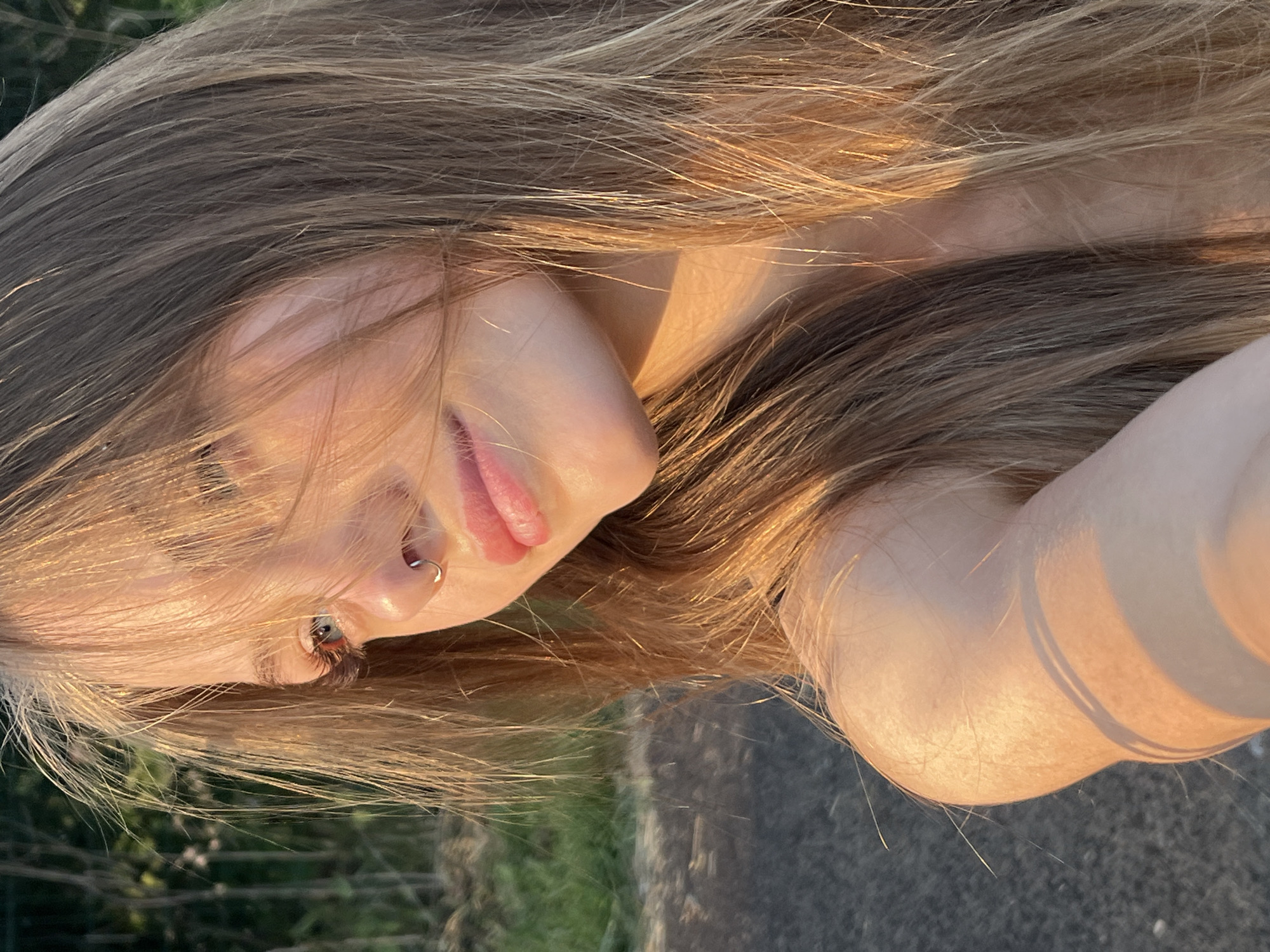 A close-up, outdoor, portrait of a young Caucasian female with long, blonde-brown hair, captured during golden hour. She is looking away from the camera to her left, with her hair partially obscuring her face. Her left eye is visible, and she is wearing a silver nose ring. Her lips are slightly parted, and she has on subtle makeup. The background is out of focus, showing greenery and a dark, paved surface, suggesting a park or a road in a natural setting. The lighting indicates a late afternoon or early evening sun. The location is specified as Фоки, Russia.