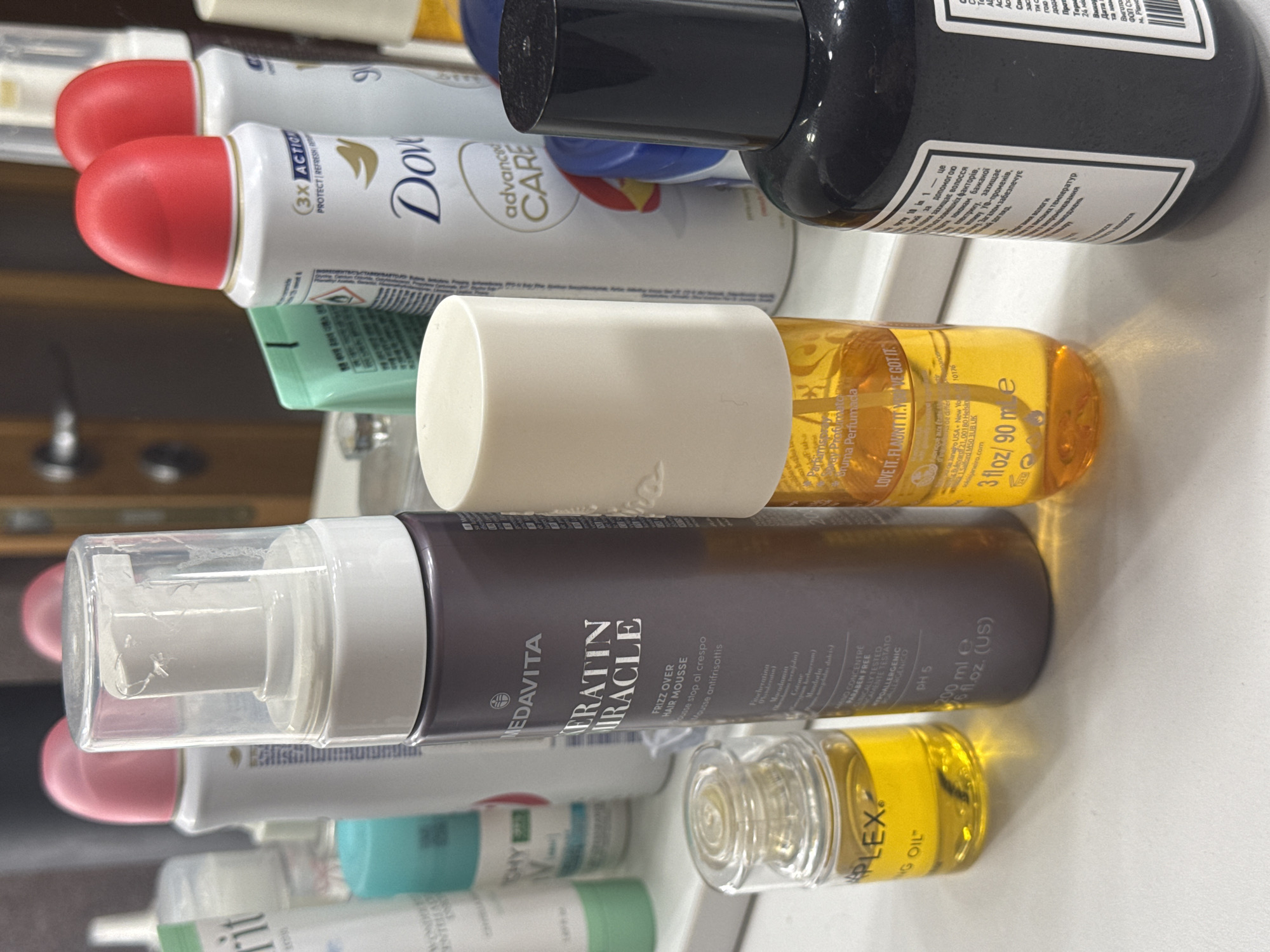 The image displays a collection of personal care products, including various bottles, cans, and tubes, arranged on a reflective surface, likely a countertop or shelf. The products are seen from a slightly elevated perspective.
Prominent items include a white Dove Advanced Care deodorant spray can with a red cap, a tall black bottle with Cyrillic text, a clear yellow bottle with a white pump dispenser labeled "LOVE IT. FLAUNT IT. WE GOT IT" and "Perfumada" (3 fl oz / 90 ml), and a dark gray/purple bottle with a white pump dispenser from MEDAVITA, identifiable as "KERATIN MIRACLE FRIZZ OVER HAIR MOUSSE" (200 ml e 6 fl.oz. (US)) with "pH 5" and "PARABEN FREE". A small clear bottle containing yellow liquid, labeled "PLEX" and "NG OIL", is also visible. Other bottles and tubes appear blurred in the background, with one partially legible as "SHY".
No specific activity is occurring; the items are stationary. The reflective surface creates a mirrored effect of the lower parts of the bottles. Notable text includes product names, sizes, ingredients like "Mandorla", and claims such as "DERMATOLOGICALLY TESTED" and "HYPOALLERGENIC". The Cyrillic text on the black bottle mentions details about hair care, protection from external factors, UV rays, moisture, and "Київ" (Kyiv) along with "Виготовл" (Manufactured) and "Дата" (Date), indicating a connection to the specified location context of Kyiv, Ukraine. The image is oriented sideways.