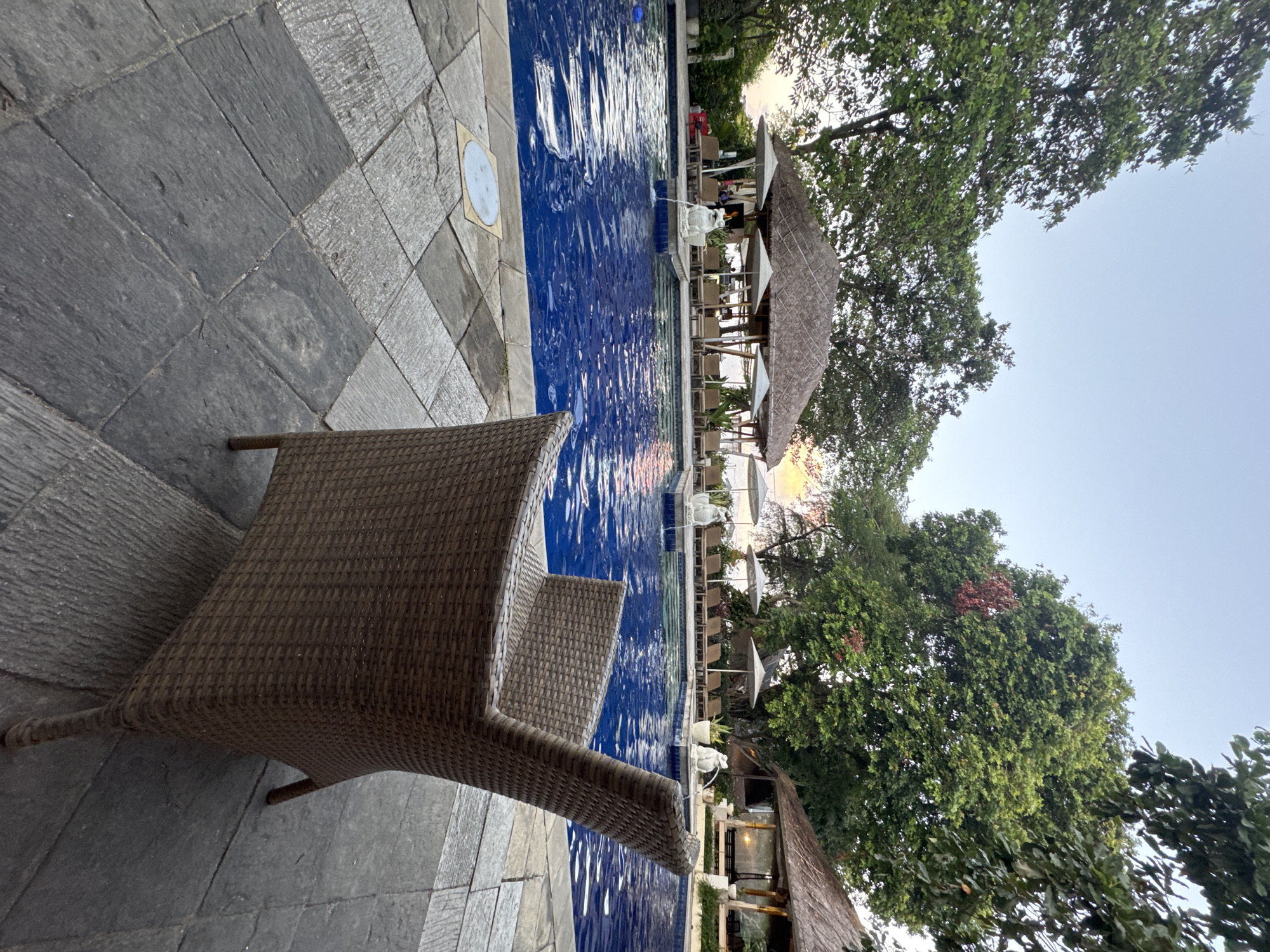 The image depicts a resort swimming pool area in Denpasar, Indonesia, during the late afternoon or early evening. The pool is large and filled with blue water, reflecting the sky. Several lounge chairs with umbrellas are visible around the pool, some shaded by thatched-roof cabanas. Two small fountains, designed as white animal figures, are placed in the pool. In the foreground, a single brown wicker chair is positioned on a stone-tiled patio. Trees with lush green foliage surround the pool area, and a glimpse of a building with a thatched roof can be seen in the background. The sky is clear with a light gradient of colors, suggesting the sun is low on the horizon. There are no people visible in the frame, implying a tranquil and unoccupied setting.