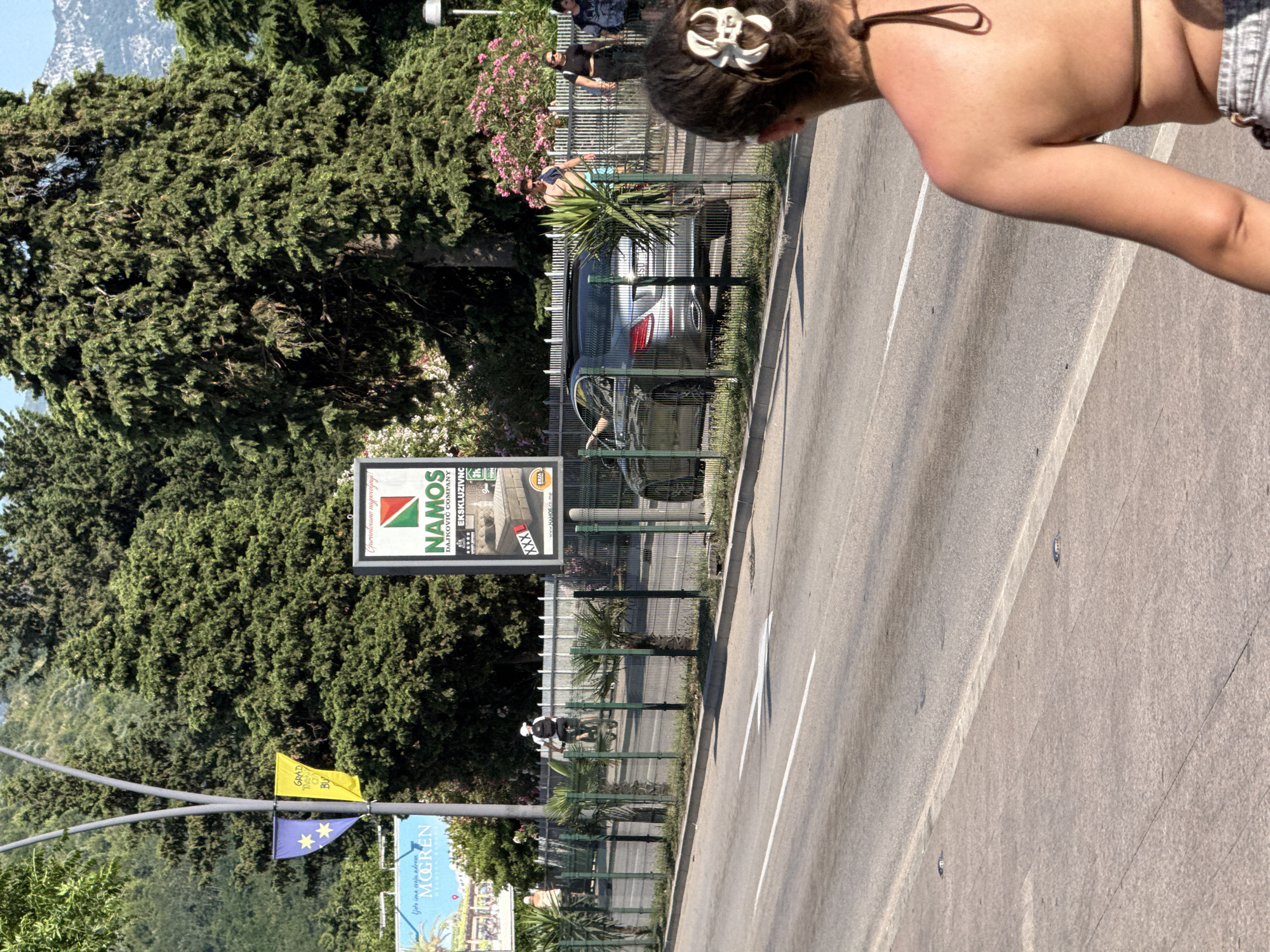 This media file is a photograph taken outdoors, likely during the daytime, with the sun casting strong shadows, suggesting a clear, sunny day. The scene appears to be a street or promenade in a coastal town, possibly Budva, Montenegro, given the visible text cues.
In the foreground, we see the upper torso of a person, facing away from the camera and to the right. They have dark hair styled with a white clip and are wearing a brown bikini top. Their arm is extended downwards, and they are positioned to the right of the frame.
The background is dominated by lush green trees, likely cypress or pine, with some pink flowers visible amongst them. A metal fence runs horizontally across the middle ground, separating the foreground from the area behind it. Through the fence, a modern silver car is parked, and a person is partially visible near it, wearing a blue top. Further down the fence, another person on a bicycle is visible, wearing a helmet.
Several signs are present. A large rectangular billboard with the text "NAMOS DAJKOVIĆ COMPANY EKSKLUZIVNC" is prominently displayed. Below it, smaller flags are hung, one yellow with text partially visible, and another blue with yellow stars, reminiscent of the European Union flag. Another blue sign with the word "MOGREN" and some text below it is also visible, along with the phrase "Ljeto ima svoju adrem".
The overall impression is a sunny day in a scenic, possibly tourist-oriented location, with a mix of natural elements and signs of modern life. The presence of the flags and the "MOGREN" sign strongly suggest a location in Montenegro, likely near the coast. The activity appears to be people going about their day, with one person cycling and a car parked nearby.