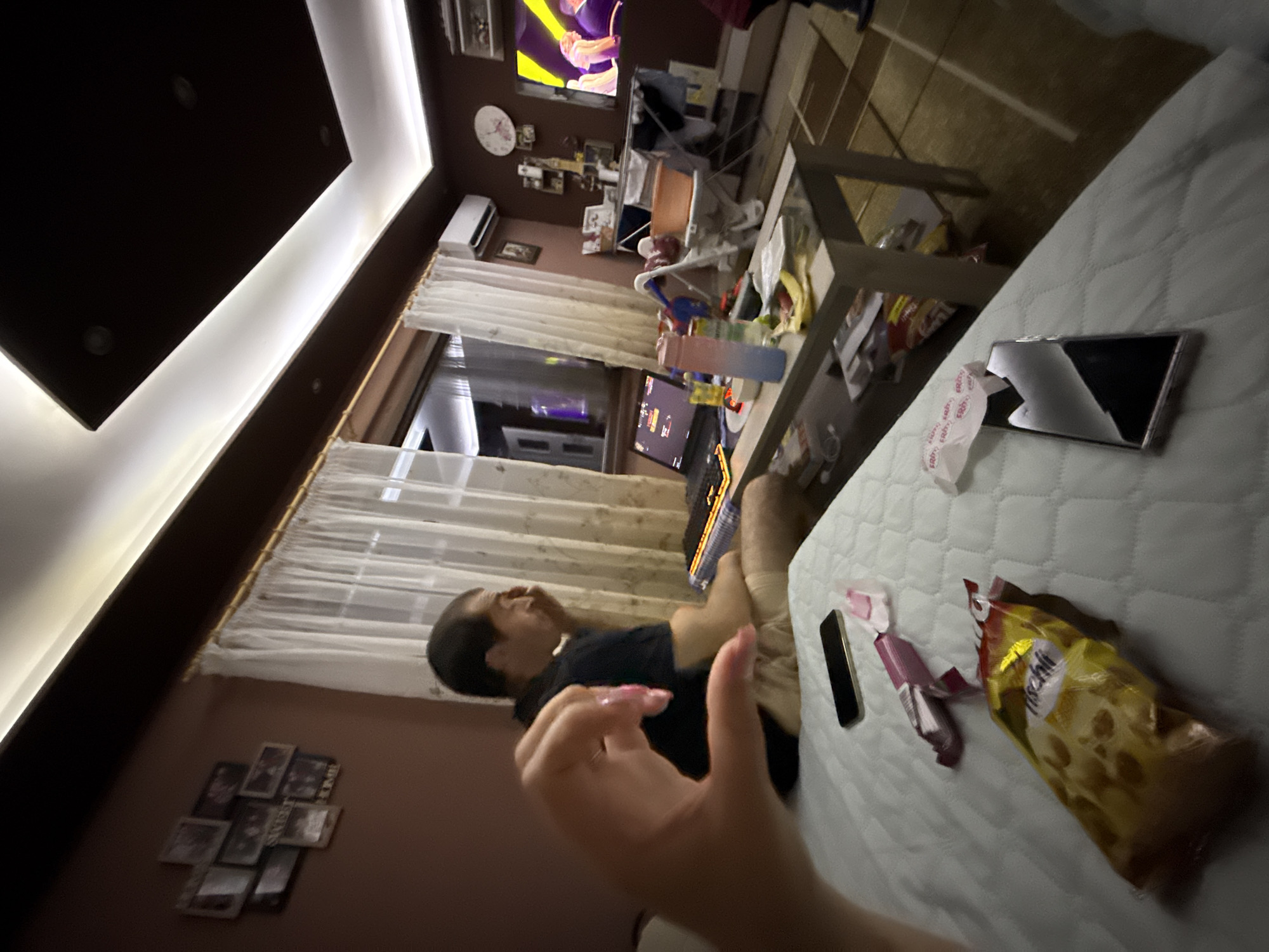 A person lies on a bed in a room. The room has a TV, a window with curtains, a ceiling light, and a wall with pictures. On the bed is a phone, a bag of chips, and some candies. A hand is raised in the foreground. The image was taken indoors and there are no visible indications of the location being in Sofia, Bulgaria.