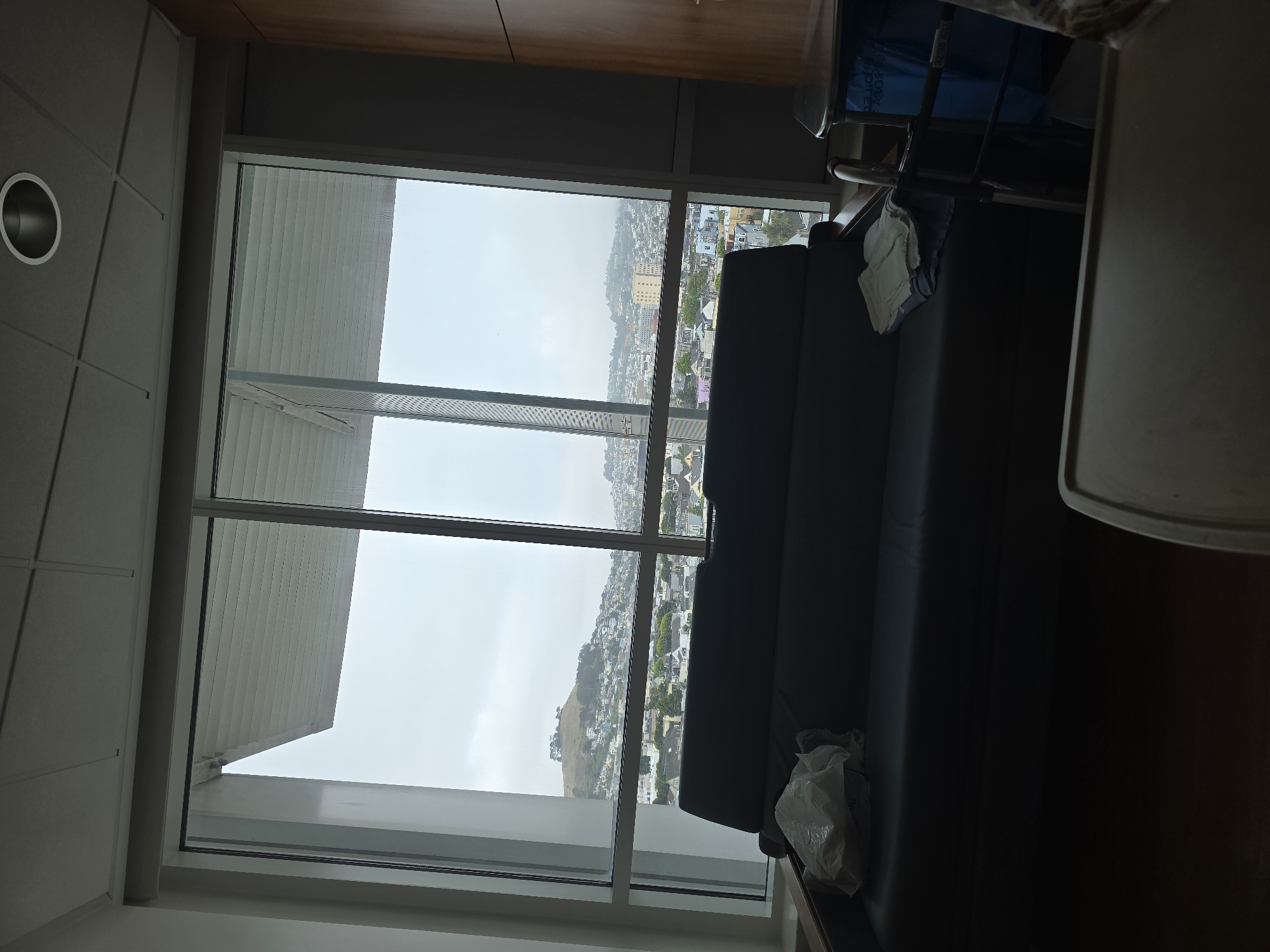 The photograph shows an interior scene, likely a hospital room, with a view of San Francisco through a large window. The window is divided into multiple panes, and the view shows a cityscape with rolling hills, buildings, and a cloudy sky. In the foreground, there is a dark-colored couch or bench with crumpled white cloth. A medical tray table is present. The walls of the room are a light gray color, and the ceiling appears to have a grid pattern. The room appears clean and relatively sterile. The view outside the window offers a glimpse of San Francisco's distinctive urban landscape, with its characteristic hills and architecture. The location is most likely Bernal Heights and Mission District, which may be inferred from the topography and urban fabric seen in the image.
