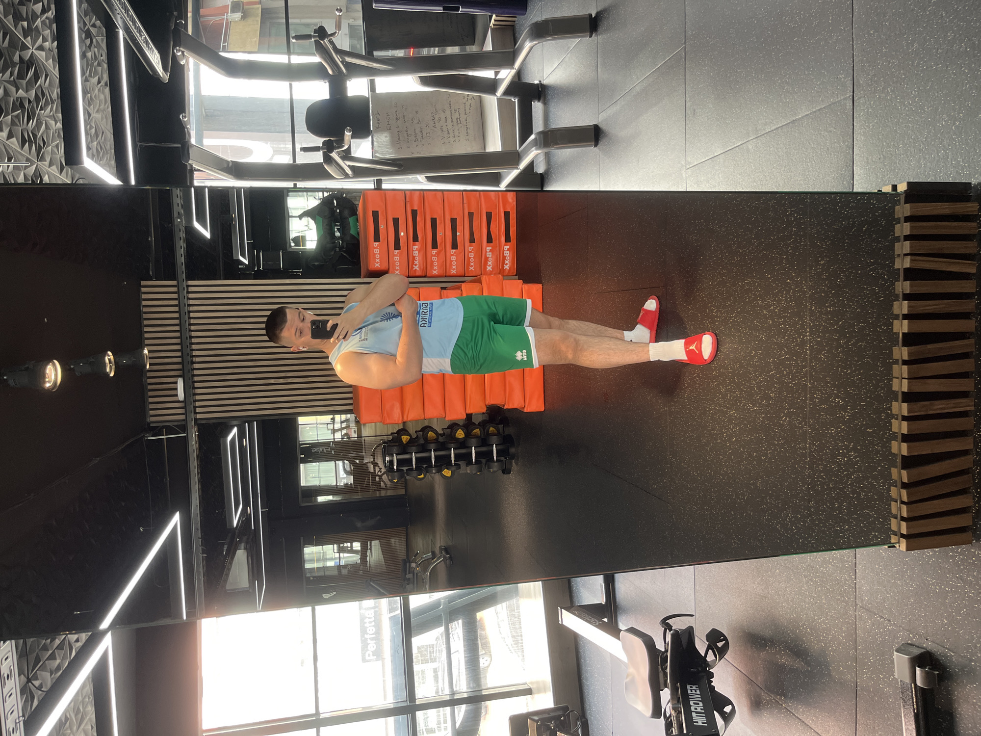 A man in a light blue tank top and green shorts, wearing white socks and red slides, is taking a selfie in a gym. He is standing in front of a large mirror that reflects various gym equipment including a rowing machine, dumbbells, and an exercise machine. The gym features black walls, light fixtures, windows showing a street view with building signage. Stacks of orange boxes are visible. Text on a nearby exercise machine is in cyrillic. The word "Perfetta" is visible in the reflection. "HIIT ROWER" is displayed on the rowing machine. The word "Sofia" and "Bulgaria" are mentioned.