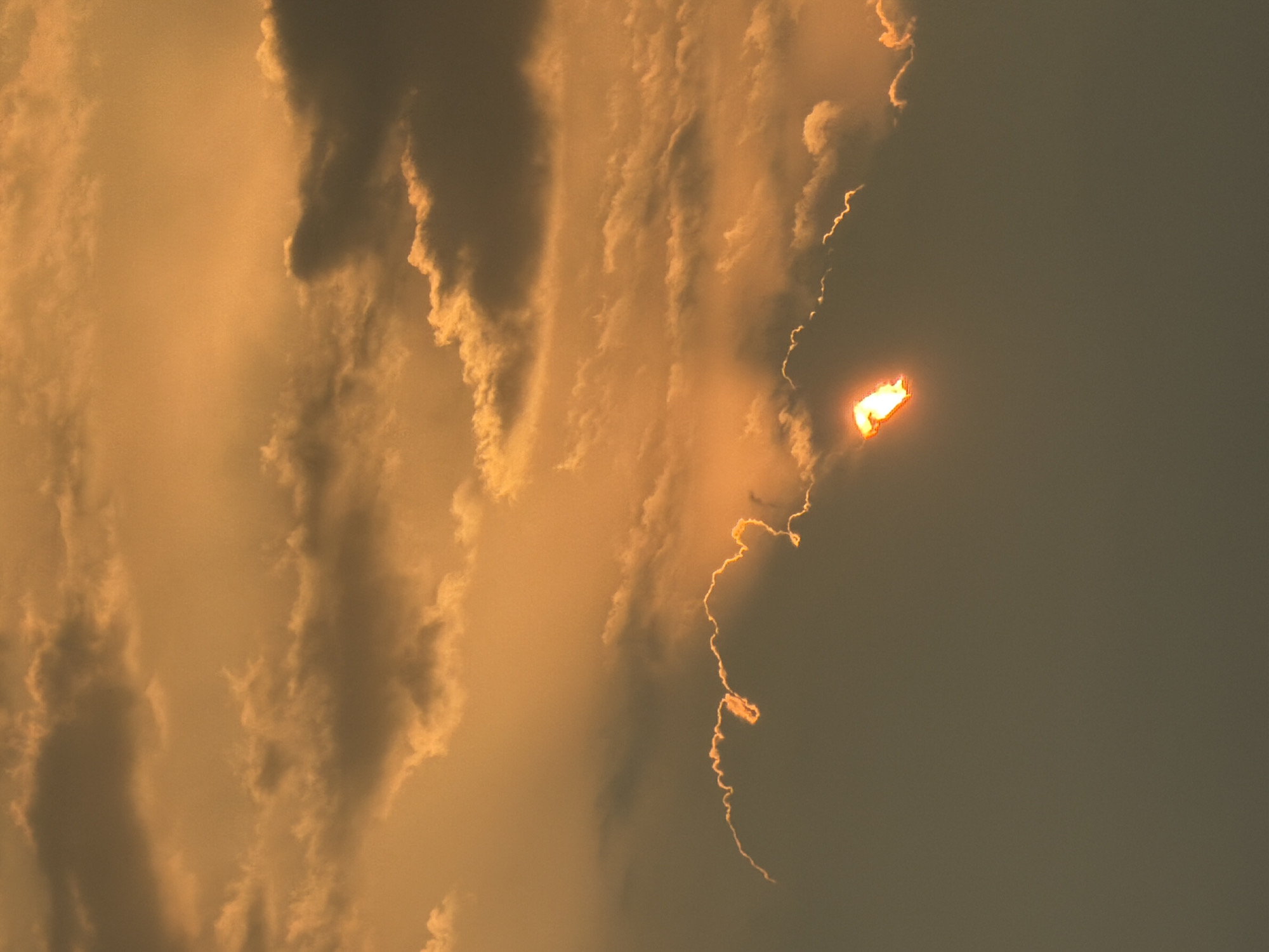 The image shows a sky, likely at sunset or sunrise, with heavy cloud cover on the left side. The sky is a mix of deep grey and orange, giving the impression of a dramatic weather event. A bright orange-yellow object that looks like fire is suspended on the right side of the image, seemingly attached to a wisp of a cloud. This object emits a tail of wispy light. The lack of recognizable buildings or landmarks prevents a definitive location identification within Cuautla, Mexico, or elsewhere. No people are visible in the image.