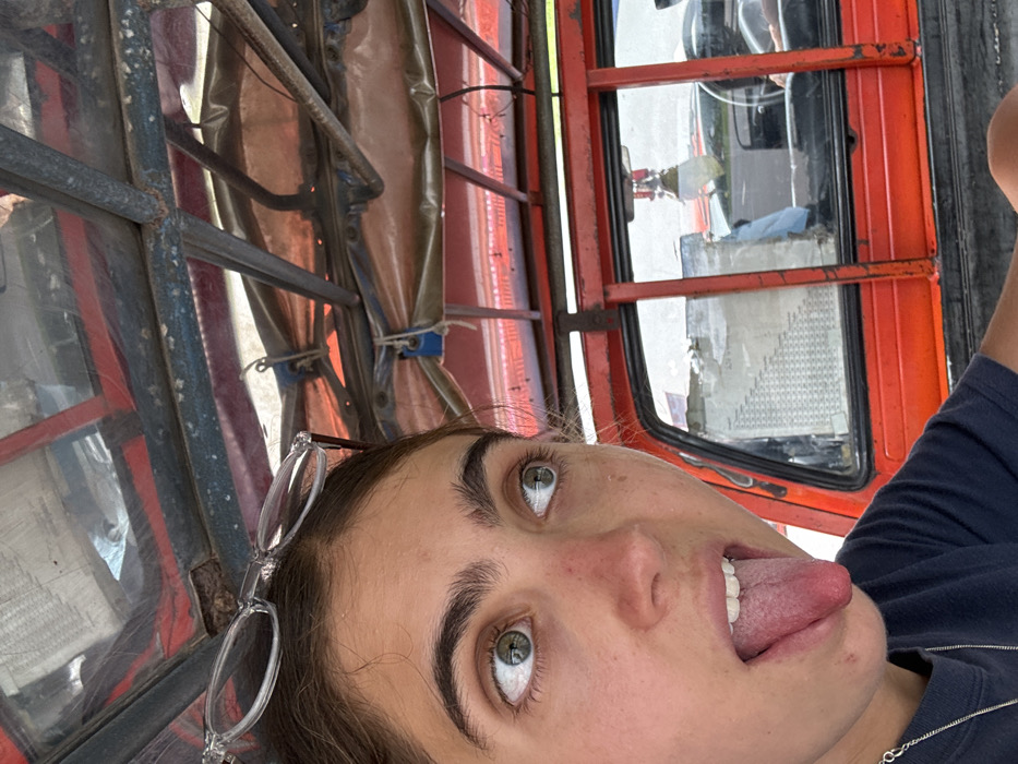 A young female is photographed in Tambon Tha Uthae, Thailand, occupying the rear passenger area of a red utility vehicle, likely a modified pickup truck or songthaew. Her fair-skinned face is partially visible in the foreground, angled upwards and to the right, with prominent dark eyebrows and green eyes looking sideways. Clear-framed glasses rest on her forehead. Her mouth is open, and her tongue is extended. She wears a dark t-shirt and a silver chain necklace.
The vehicle's interior features a red metal frame structure and an orange-red tarp canopy secured by blue ropes overhead. Behind the subject, a red-painted metal partition and a rear window are visible. Through this window, a light-colored document or chart is taped, and reflections reveal part of a driver's cabin, including a steering wheel and a hanging white air freshener. Exterior elements visible through the window suggest another vehicle and an outdoor environment, possibly a road. The lighting indicates daylight conditions.