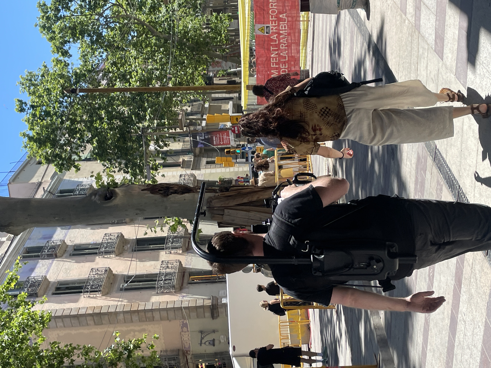 A man wearing black clothing stands with a camera support system on his back. A woman with long, curly brown hair walks in front of him, wearing a patterned tan top and light tan pants. They are on a pedestrian walkway, possibly La Rambla in Barcelona, Spain, given the banner reading "M fent la reforma de la Rambla" (We are doing the reform of La Rambla). The architecture in the background includes buildings with multiple floors and decorative ironwork on the balconies. Green trees provide shade, and the sky is blue. A white truck is visible behind the man, and yellow crowd control barriers are nearby. Other pedestrians are also present in the background.