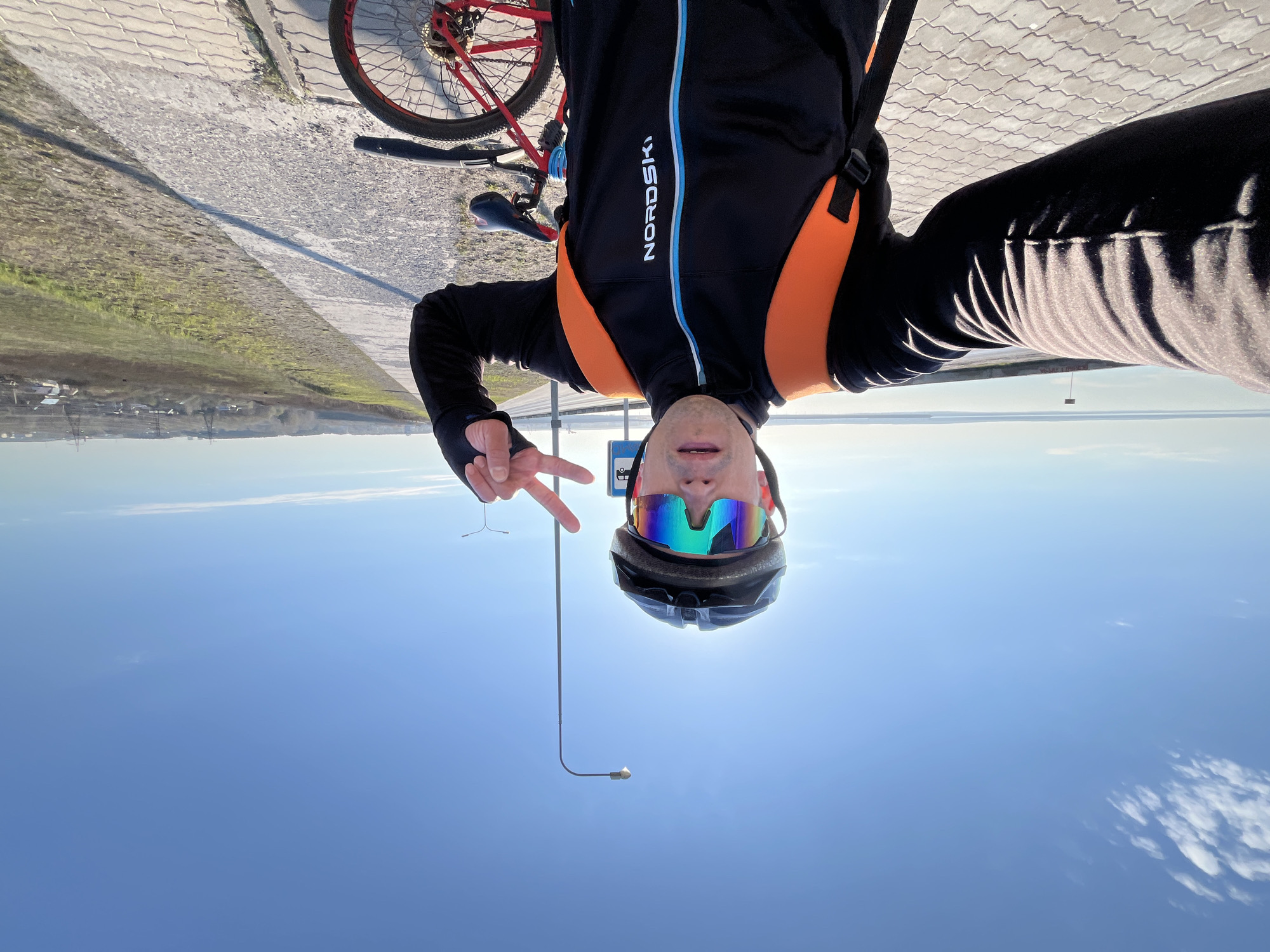 A man wearing a black and orange Nordski jacket, helmet, and multicolored sunglasses poses for a selfie with a peace sign. A red bicycle is partially visible in the top left corner. The background shows a paved area, a landscape with water, and a bright blue sky with some clouds. A street lamp is visible below the man. This photo was taken in the city of Chaikovsky, Russia.