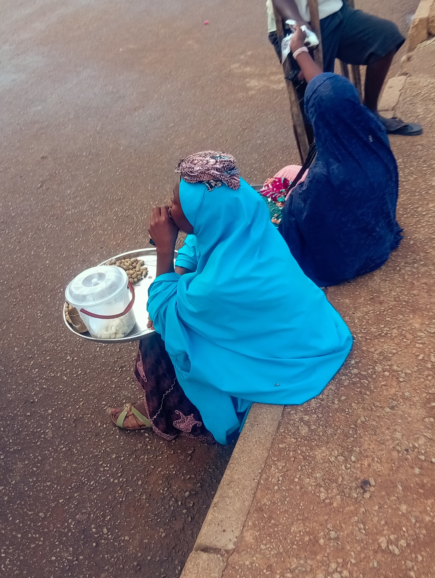 The image shows a street scene in Bukuru, Nigeria. Two women are visible, one in the foreground and another in the background. The woman in the foreground is wearing a bright blue hijab and a patterned skirt. She is seated on the ground with a metal tray in front of her. On the tray are a white bucket with a red handle, filled with what appears to be chopped vegetables, and a pile of peanuts. She is holding something to her mouth, possibly eating. Her sandal is visible on her right foot.
The second woman is in the background, wearing a dark blue hijab. She is standing next to a wooden structure, possibly a market stall or a bench, and appears to be interacting with a person whose legs are visible standing behind her. This person is wearing dark shorts and dark shoes. The ground is a mix of asphalt and a rough, reddish-brown dirt surface. The lighting suggests it is daytime, possibly overcast given the diffused light. The overall scene depicts a common street vending or informal market activity. There is no visible text in the image.