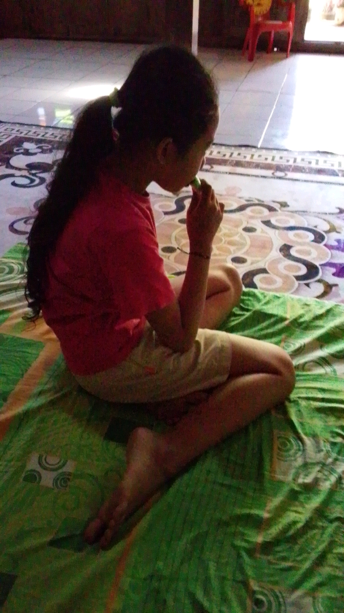 A young girl with dark, long hair tied in a ponytail is sitting cross-legged on a green and white patterned blanket. She is wearing a pink t-shirt and light-colored shorts. Her right hand is raised to her mouth, and she appears to be eating something green, possibly a small vegetable or a piece of candy. The girl is facing away from the camera, and her profile is visible. Her left leg is bent and resting on the blanket, while her right leg is tucked beneath her. Her bare feet are visible at the bottom of the image, with her toes pointed towards the left. The background shows a tiled floor with a patterned rug and a glimpse of a red chair and a bright window with sunlight streaming in. This scene is set in Lawu, Indonesia.