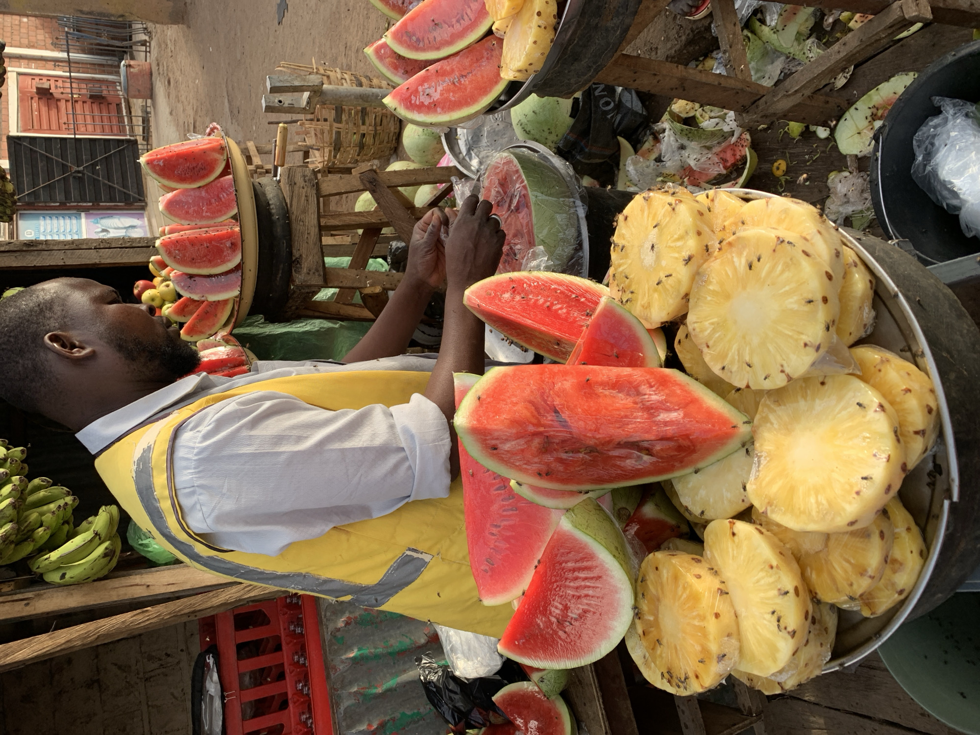 A vendor in Jos, Nigeria, stands behind a cart piled with fresh-cut fruit. The vendor, a dark-skinned man with a beard, wears a white shirt and a yellow safety vest with grey reflective stripes. His hands are occupied with slicing a watermelon. The cart is loaded with watermelon slices, pineapple chunks, and whole watermelons, some wrapped in plastic. Flies are visible on the cut fruit. A separate display holds vertical watermelon slices. To the left, a bunch of green bananas rests on a wooden surface. In the background, a building with barred windows is visible. Trash and discarded fruit rinds are scattered near the cart.