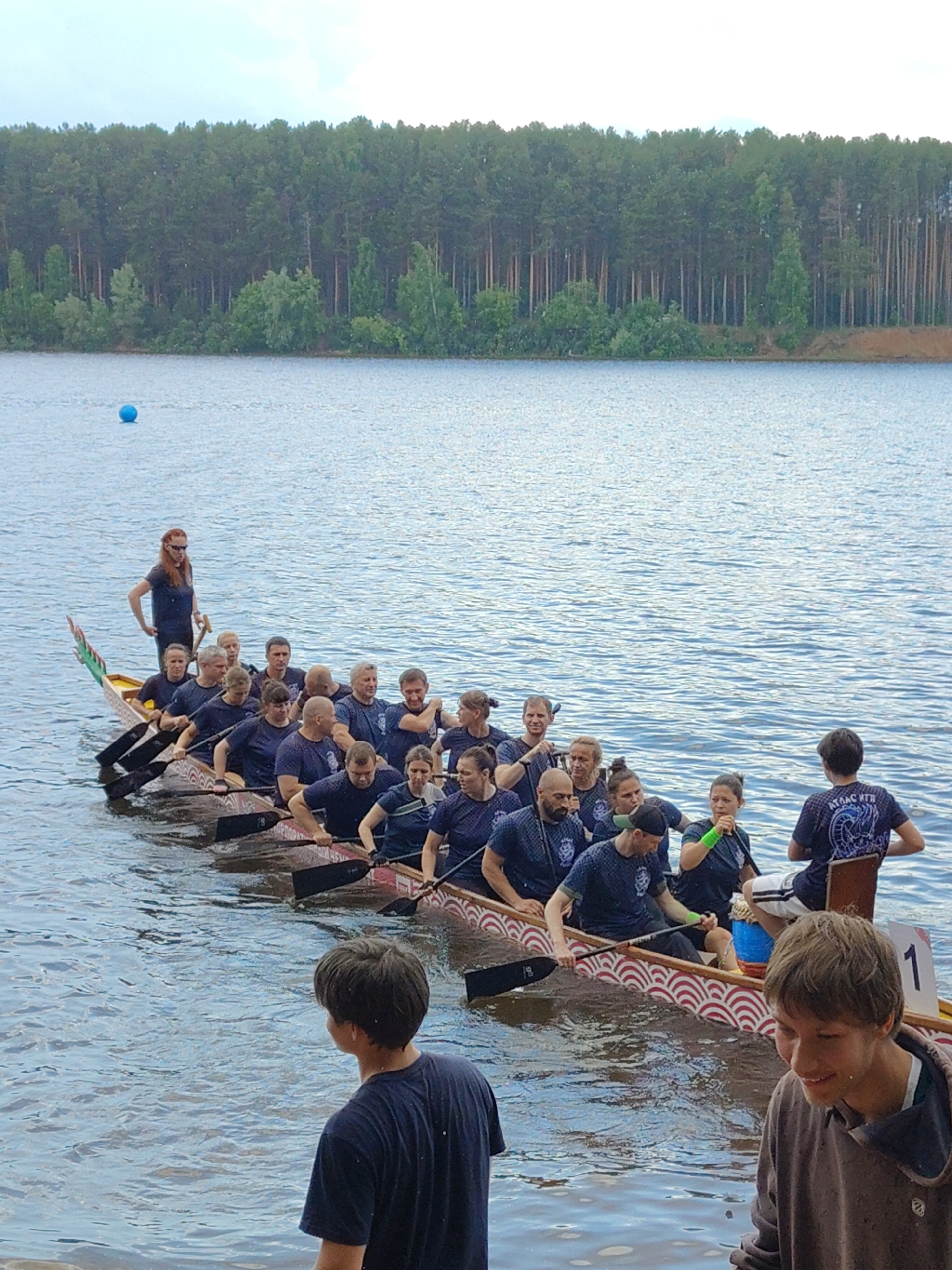 In the city of Unknown, Unknown, on a body of water, a dragon boat team prepares to paddle. The boat, decorated with a dragon's head and tail, is filled with approximately fifteen individuals wearing dark blue shirts and athletic wear. Most are seated and holding paddles, positioned for synchronized rowing. A woman with red hair stands at the stern, facing the rowers and holding a paddle upright, likely acting as the coxswain. In the foreground, two young men are visible at the water's edge, one looking out at the boat. The water is calm with a slight ripple, and a single blue buoy is visible in the distance. A dense forest lines the background, with a clear sky above.