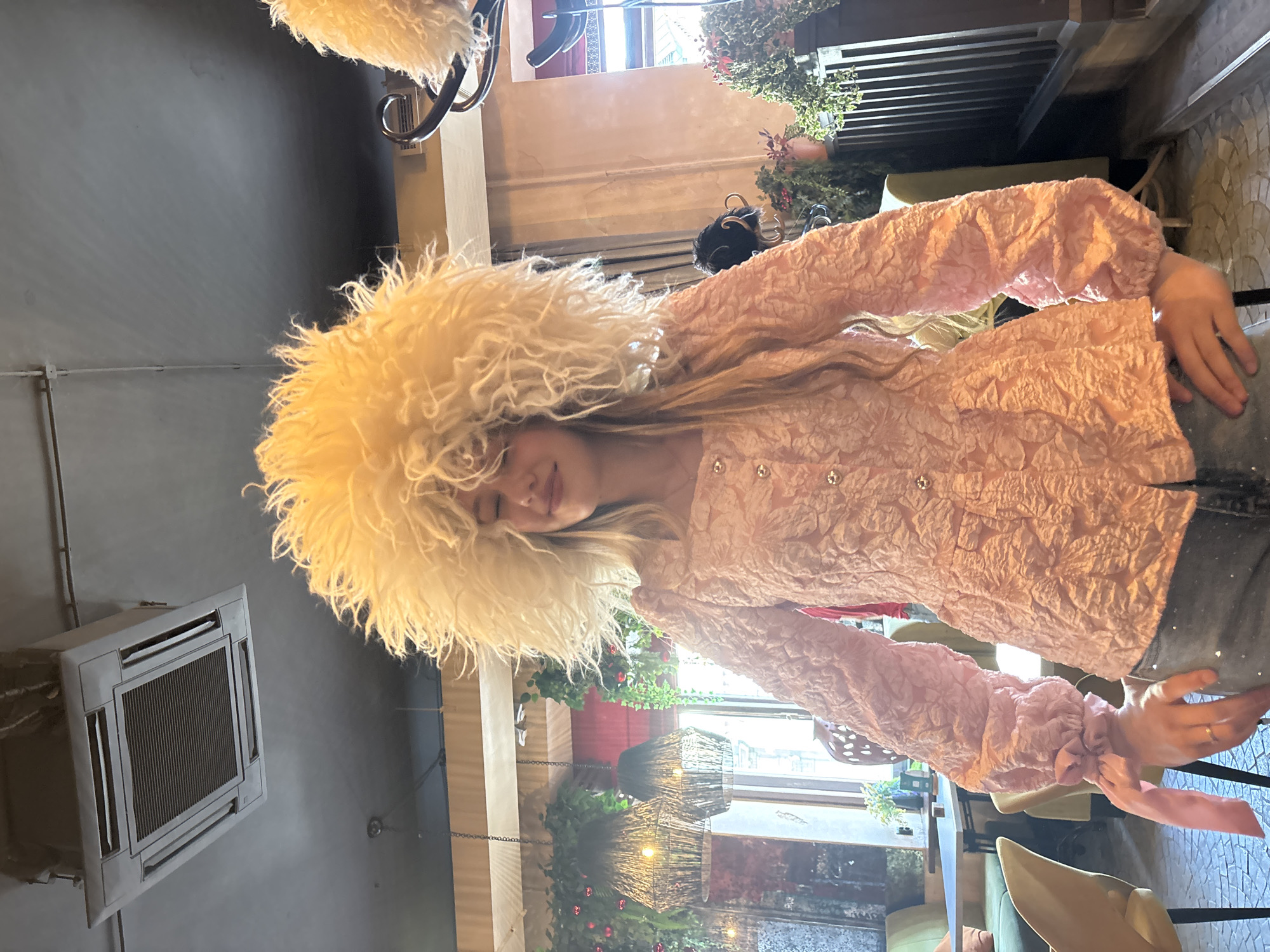 The image depicts a single female subject positioned centrally in an indoor establishment, identified as being in Saint Petersburg, Russia. Her eyes are closed, and she exhibits a slight smile, with her head subtly tilted. She wears a prominent, voluminous, shaggy hat of a light cream or pale yellow color, characteristic of a papakha-style headwear; long, light-colored hair is visible beneath the hat. She is dressed in a light pink, long-sleeved blouse featuring a textured, possibly floral, embossed pattern. The blouse has gold-toned buttons down the front and voluminous sleeves gathered at the wrists with self-fabric ribbons. She wears dark wash denim trousers with subtle light-colored embellishments. A ring is present on her left ring finger.
The ceiling is dark gray, featuring an exposed cassette-style air conditioning unit and visible utility conduits. Multiple woven pendant lamps provide warm illumination for abundant hanging foliage interspersed with red berry accents. To the left, light-colored chairs and green booth seating are arranged around tables. On the right, a warm-toned wall section features a dark wooden balustrade and a coat rack from which a smaller, dark fur hat hangs. The floor is surfaced with a mosaic pattern of irregularly shaped tiles. The overall environment suggests an upscale cafe or restaurant with an eclectic, botanical-themed decor.