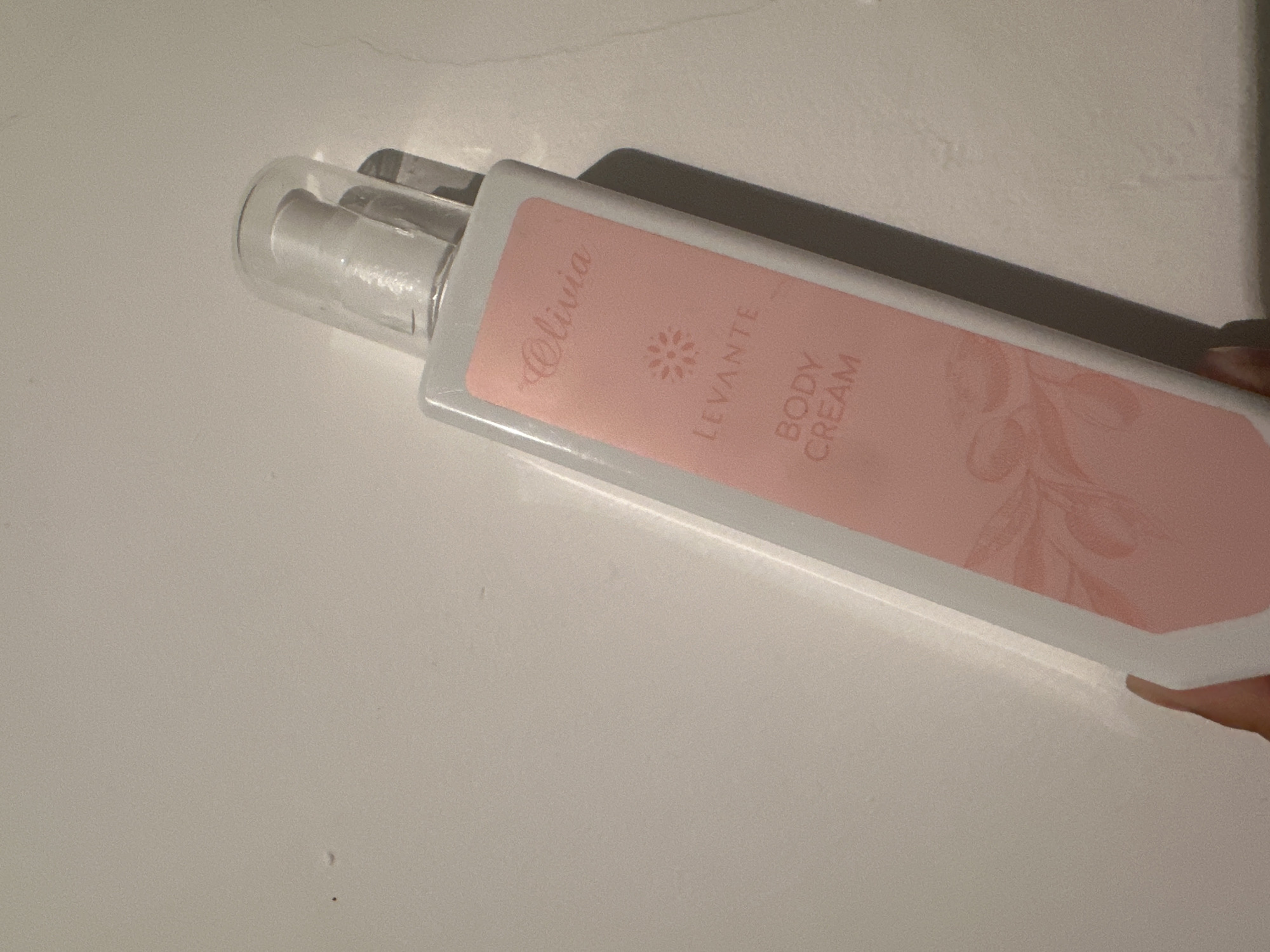 The image shows a hand holding a white bottle of "Olivia Levante Body Cream" against a plain white background. The bottle has a light pink label with floral and leaf motifs. The lighting in the image is diffuse, creating soft shadows. The exact location in Тайгараев, Kyrgyzstan, is not discernible from the image.
