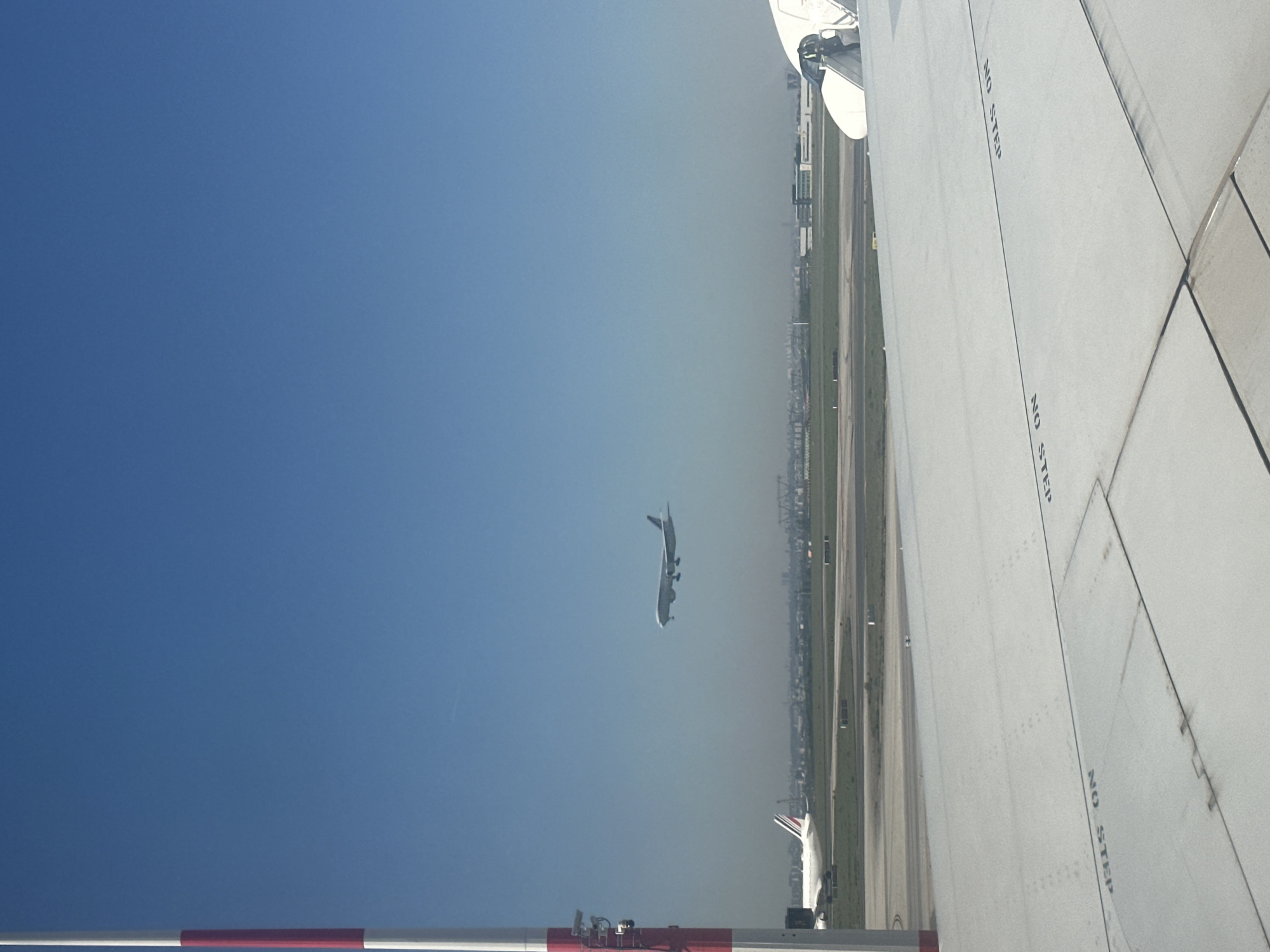 This daytime image, taken from what appears to be an airplane window or an elevated airport structure, depicts a bustling airport scene under a clear blue sky, with a hazy horizon line in the distance.
In the foreground, the clean white surface of a large airplane wing occupies the right side of the frame. Multiple "NO STEP" warnings are stenciled clearly on the wing. At the bottom of the image, a red and white striped barrier or pole is partially visible.
A prominent subject in the mid-ground is another large airplane, airborne against the blue sky, suggesting it is either taking off or landing. Its underside is visible as it ascends or descends. Further towards the lower center of the image, the white tail fin of a third airplane can be seen parked on the tarmac.
The background features an extensive airport environment with runways and taxiways, stretching towards a distant urban or industrial landscape with buildings and structures, somewhat obscured by the haze. The clear weather and visible aircraft activity indicate a busy operational day at the airport. This scene is consistent with the location context of Tremblay-en-France, home to a major international airport.