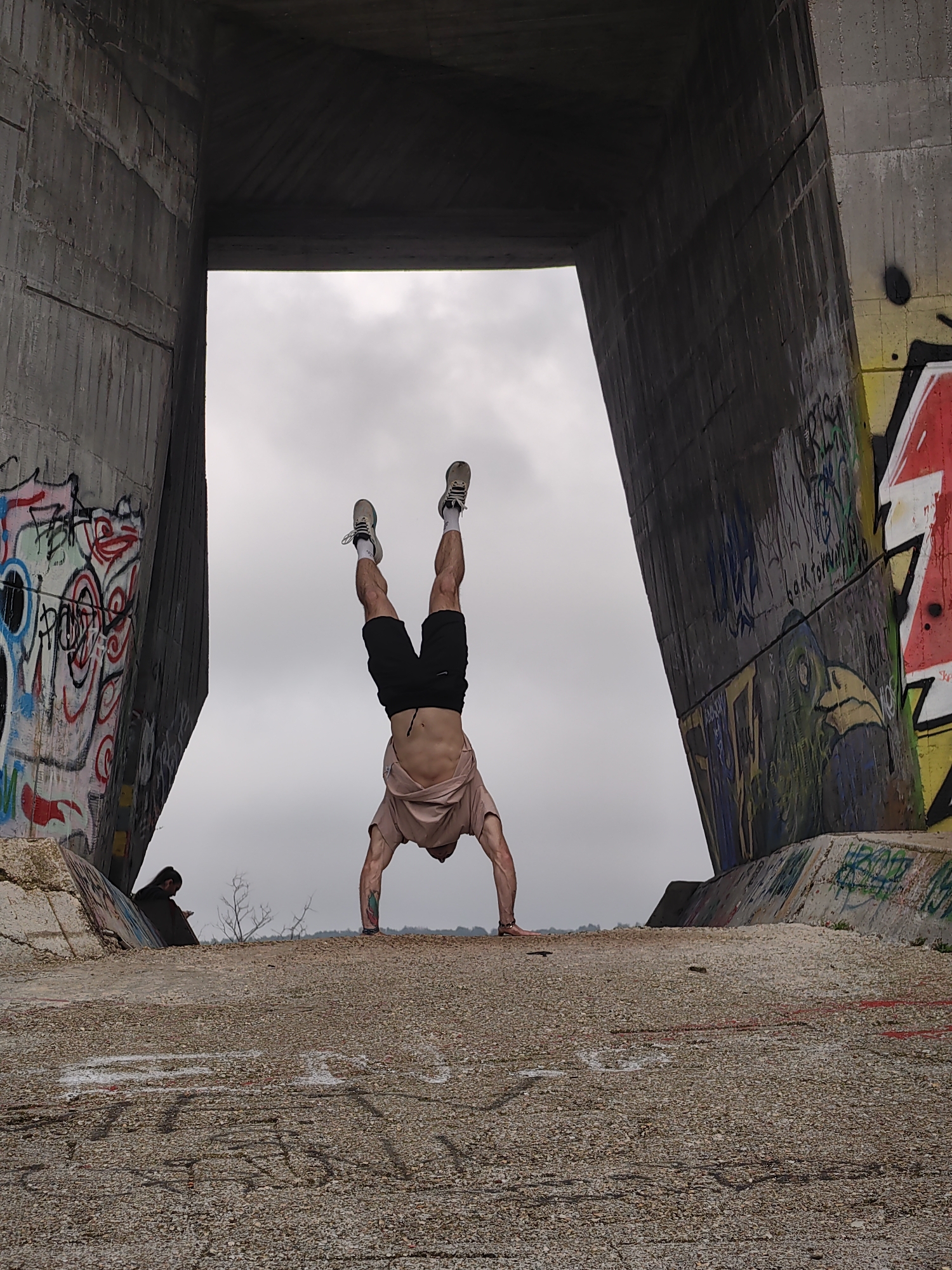 A man is performing a handstand on a concrete structure, possibly a bridge or overpass, in Varna, Bulgaria. The structure features graffiti on the support pillars. The man is wearing black shorts, a light-colored t-shirt, socks, and sneakers. Another person is sitting to the left of the standing man, with their head bowed and looking at a device. The background is a cloudy sky.