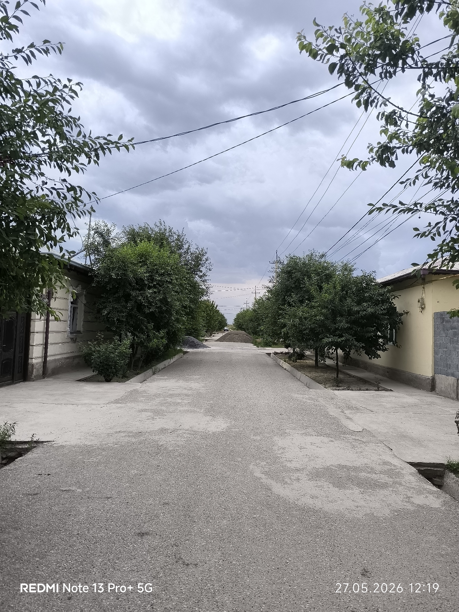 The image depicts a street in Pakhtakor, Uzbekistan. The street is lined with single-story residential buildings on either side, separated by trees and vegetation. The street surface is paved with a light-colored material, possibly gravel or asphalt. The sky is overcast with gray clouds. Electrical wires are strung overhead. No people are visible in the image. In the distance, a pile of gravel or construction material is present. The photograph was taken with a Redmi Note 13 Pro+ 5G, with the date and time stamp indicating 27.05.2026 12:19.