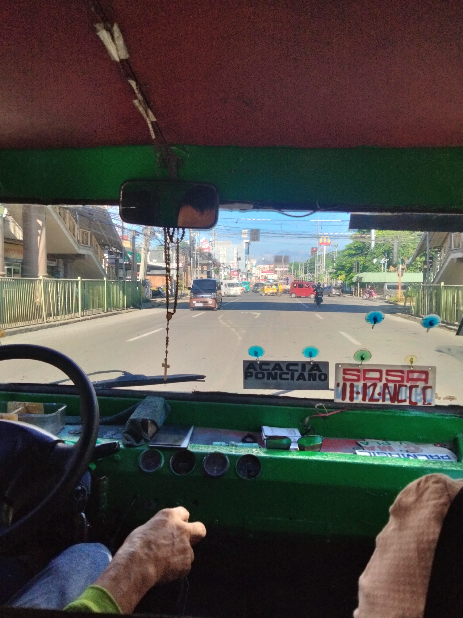 The image is taken from inside a public transportation vehicle, likely a jeepney, in Davao City, Philippines. The view is looking out the front windshield. A man's hands are visible on the steering wheel and gear shift. The dashboard is green with various gauges. Signs reading "ACACIA PONCIANO" and "SOSA 11-12-NCCC" are attached to the windshield. A rosary hangs from the rearview mirror. Outside, the street is lined with buildings and pedestrian overpasses. Traffic includes cars, motorcycles, and other jeepneys. The time of day appears to be morning or early afternoon, based on the lighting.