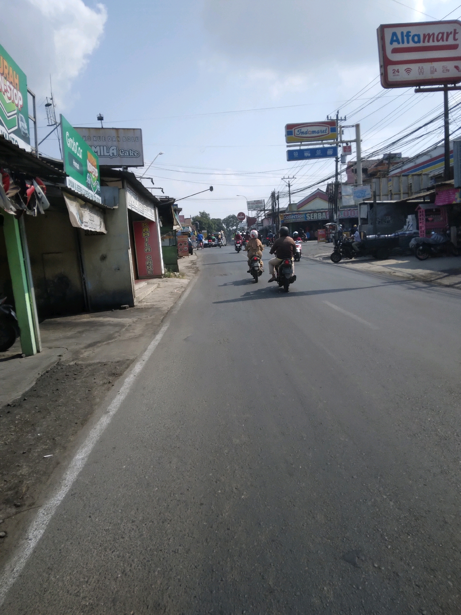 The image captures a street scene in Sokaraja, Indonesia, under a bright, partly cloudy sky. The perspective is from a vehicle traveling on an asphalt road, with the right side of the road featuring a sidewalk and shops. Several signs are prominently displayed, including a large "Alfamart" sign and an "Indomaret" sign, indicating the presence of convenience stores. Other signs suggest businesses offering cakes and possibly food.
Several people are present, primarily riding motorcycles. Two individuals are clearly visible on motorcycles in the foreground, heading away from the camera. Further down the road, more motorcycles and people can be seen, indicating active traffic. The buildings lining the street appear to be a mix of commercial establishments, some with open fronts or visible merchandise. Electrical wires are strung across the street, a common sight in urban Indonesian settings. The overall atmosphere suggests a bustling commercial area with ongoing daily activities.
