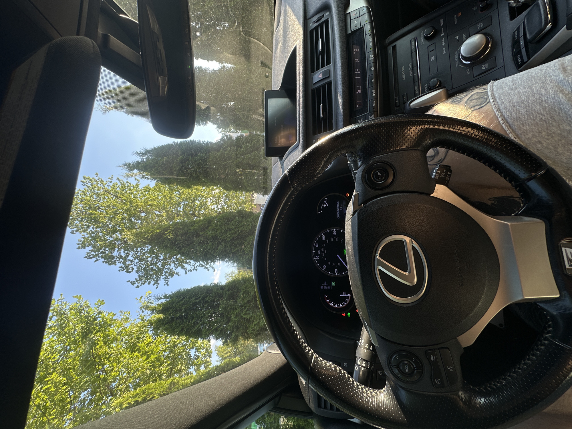 The image shows the interior of a Lexus car, viewed from the driver's perspective. The steering wheel, dashboard, and center console are prominent. The steering wheel has a Lexus logo in the center and several control buttons. The dashboard displays a speedometer and other gauges, indicating the car is in motion. The center console features a display screen, climate controls, and audio system buttons, including options for "RADIO," "MEDIA," "ECO," and "SPORT."
Through the windshield and side windows, a bright, sunny outdoor scene is visible, with lush green trees and a clear blue sky. The sunlight suggests it is daytime. The driver's leg, with tattoos visible, is also partially in the frame, indicating someone is driving.
Visible text includes brand names like "LEXUS," and function labels on the car's controls such as "AUTO," "OFF," "MODE," "CRUISING RANGE," "DISP," "RES," "SET," "RADIO," "MEDIA," "ECO," and "SPORT." The climate control displays show "71" and "72," likely representing temperature settings in Fahrenheit. The speedometer shows a speed of approximately 20-40 mph.
The overall impression is that of a person driving a comfortable, modern car on a sunny day through a tree-lined area. The setting appears to be a suburban or rural road, given the abundant greenery.