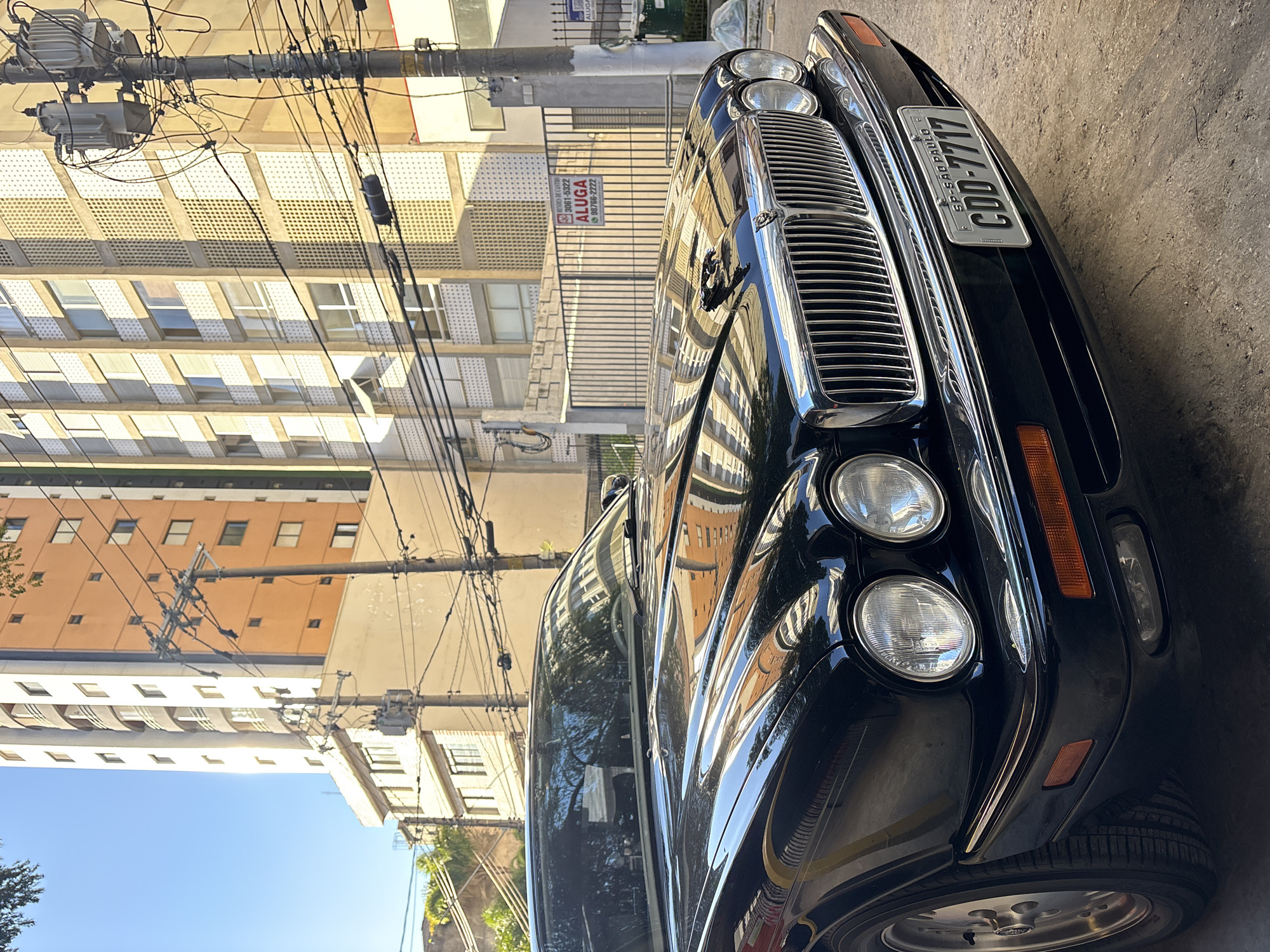 A black Jaguar XJ is parked on a street in São Paulo, Brazil, during the day. The car is in the foreground, facing slightly to the right. The building behind it is a tall, multi-story structure with an orange and white facade, featuring numerous windows. Several utility poles with a dense network of wires are visible in front of the building. A metal fence with a sign reading "ALUGA" (rent in Portuguese) is also present. The sky is clear and blue, indicating sunny weather. The street appears to be paved with asphalt and gravel. There are no people visible in the image.