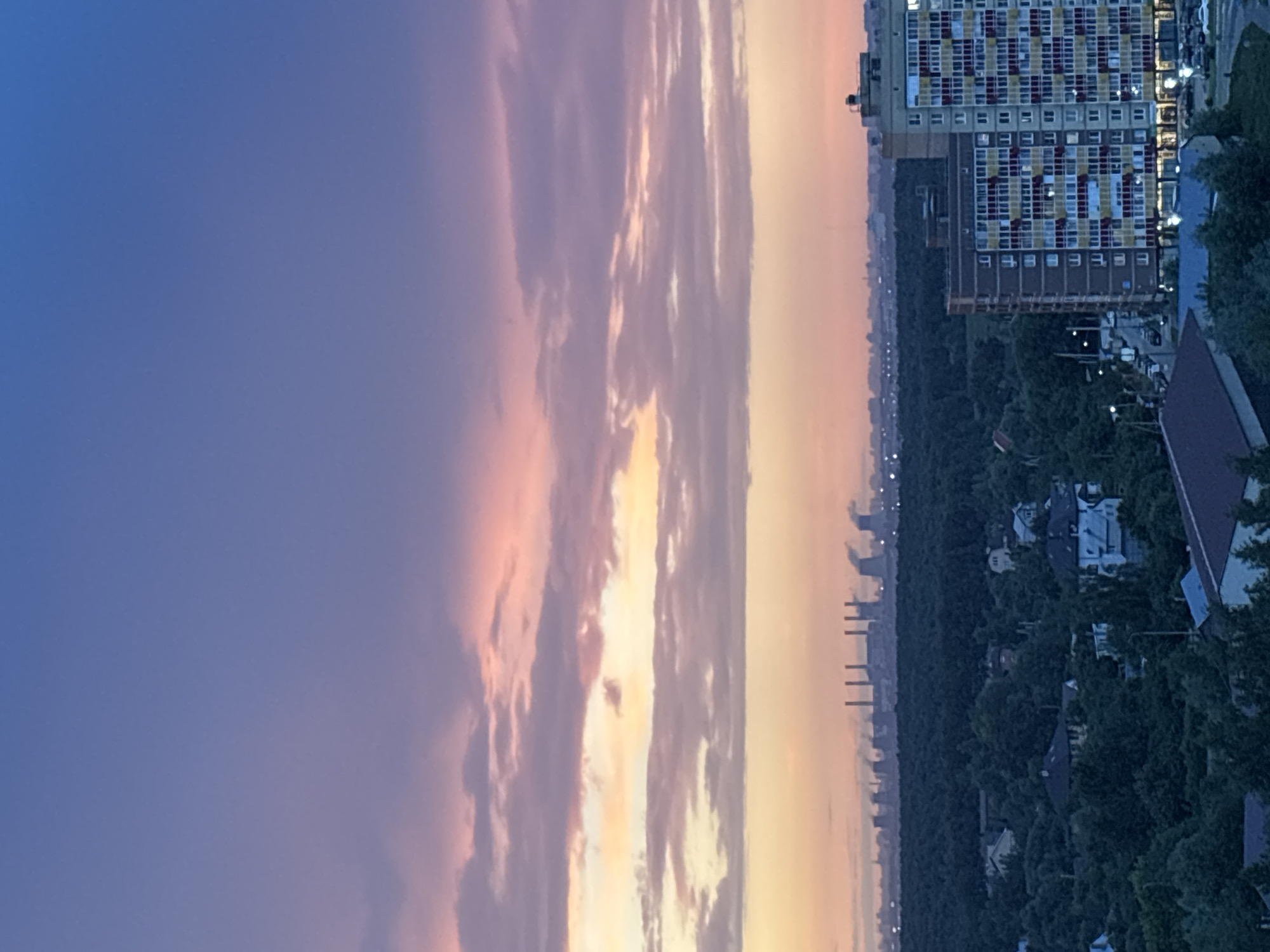 This image captures a twilight scene overlooking Misaylovo, Russia. In the foreground, a modern, multi-story apartment building with a colorful facade of yellow, red, and blue panels stands prominently. Below the apartment building, a dark, dense forest stretches across the middle ground, interspersed with scattered houses and trees. In the far distance, a line of industrial smokestacks emits plumes of smoke against the soft hues of the sunset sky. The sky itself transitions from a deep blue at the top to soft shades of pink and yellow closer to the horizon, indicating the sun has recently set. The overall impression is a juxtaposition of urban development, nature, and industry.