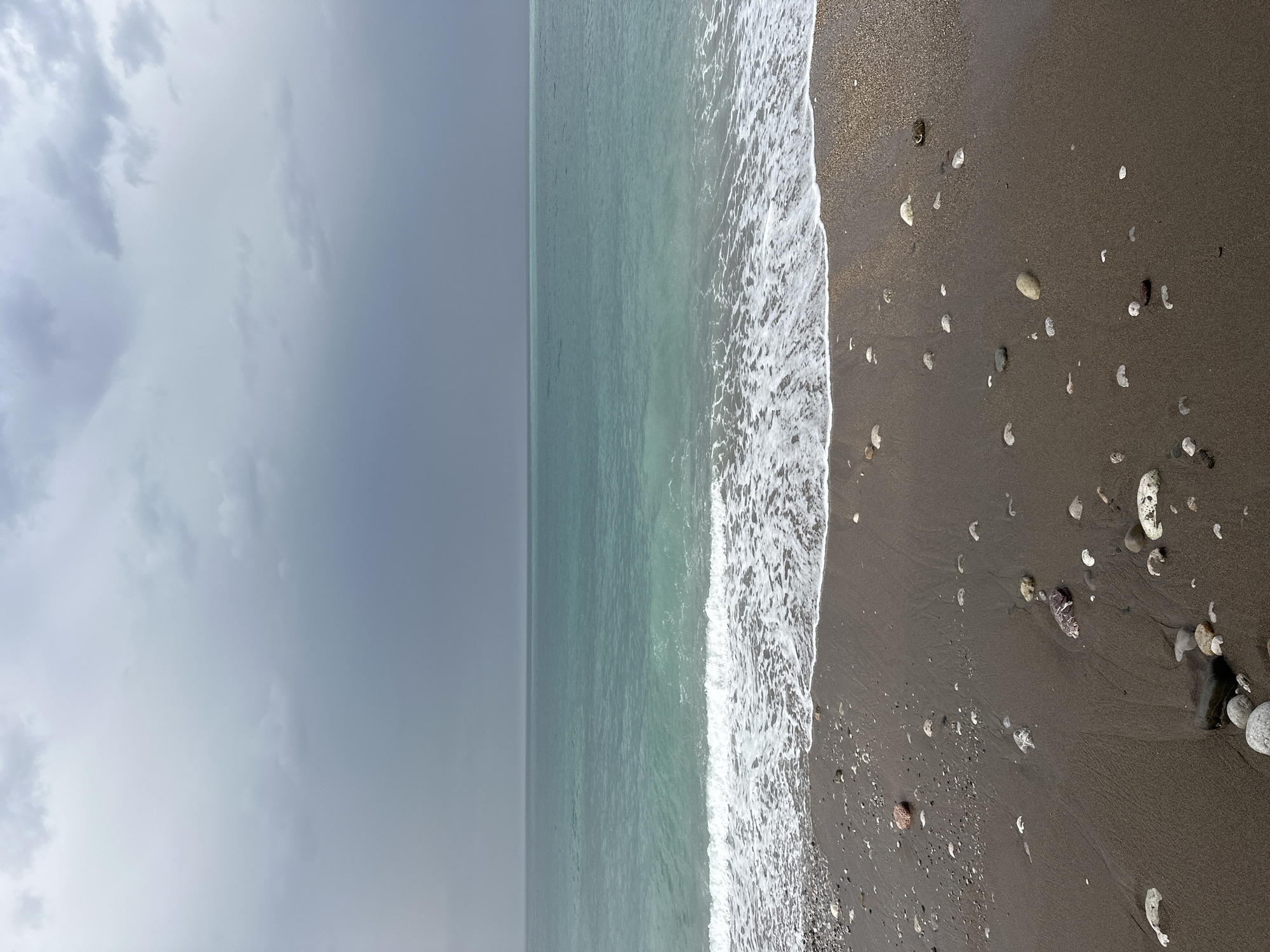 The image shows a beach scene on a cloudy day. The left side of the image shows a sky filled with gray, overcast clouds. Adjacent to the sky is the ocean, its surface a muted, light blue-green color. White foamy waves are visible where the ocean meets the shore. The right side of the image depicts the dark sand or pebble beach, scattered with rocks of various sizes and colors. This scene is located in the city of Bar, Montenegro.