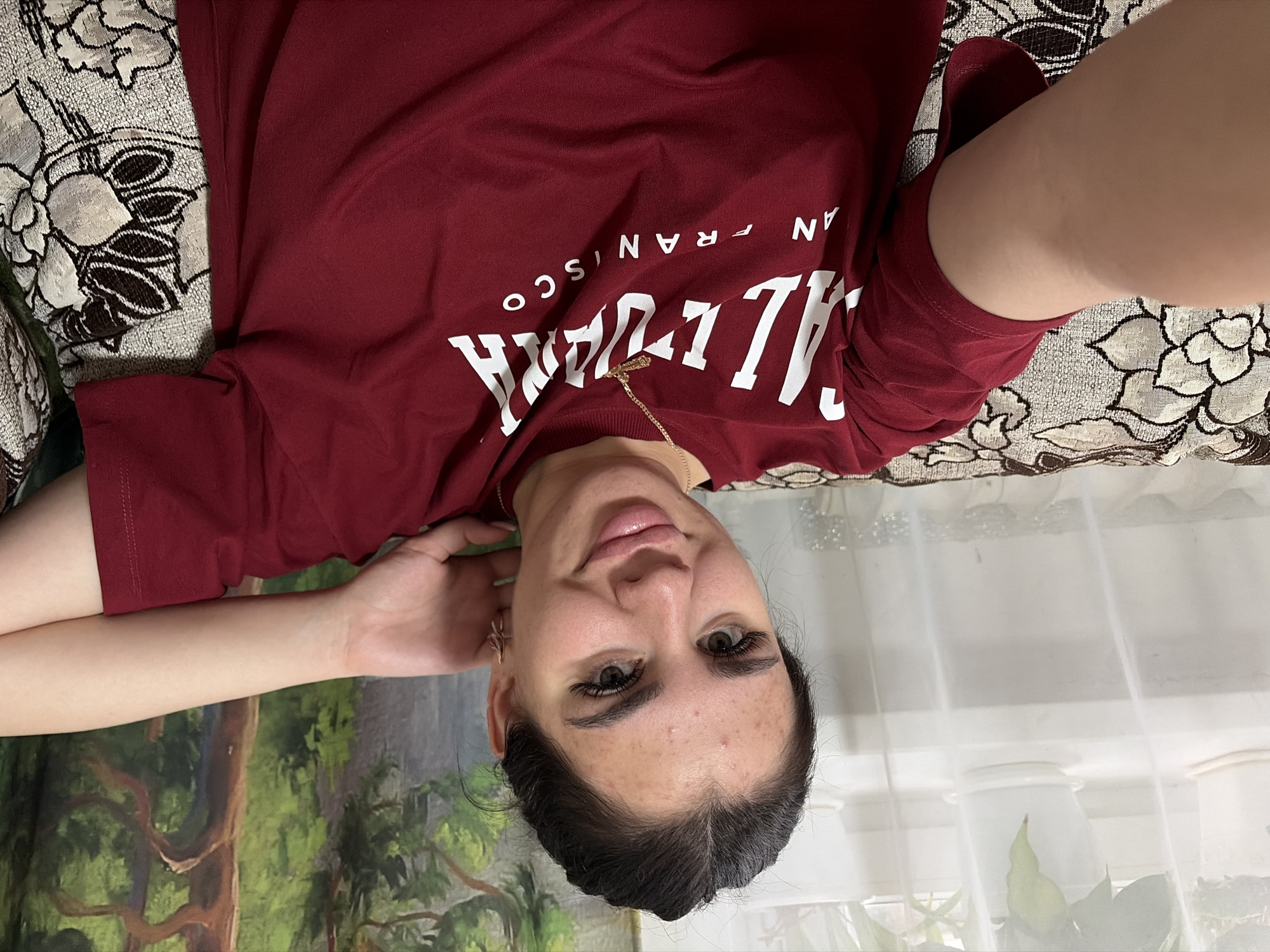 A young woman with dark hair pulled back and notable makeup, including false eyelashes, is pictured in a close-up selfie. She is wearing a maroon t-shirt with white lettering that appears to read "CALIFORNIA SAN FRANCISCO." She is seated on a sofa with a floral patterned upholstery. Her left arm is bent behind her head. The background includes a curtain and a painting or tapestry depicting trees and greenery. There is no identifiable information indicating the location is in Фоки, Russia.