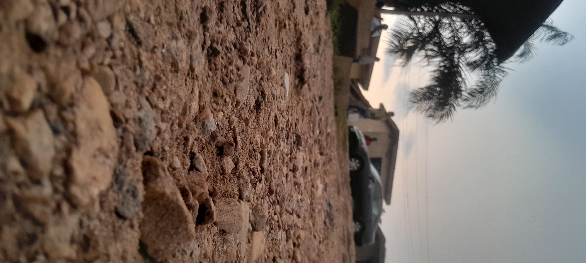 The image depicts a ground-level perspective of a dusty, unpaved area in Abuja, Nigeria. The foreground is dominated by loose soil and gravel. In the middle ground, several people are visible standing near some low-lying buildings. A dark-colored sedan is parked to the right of the buildings. In the background, the sky is overcast, suggesting it might be late afternoon or early evening. A palm tree is partially visible on the left side of the frame, its fronds blurred due to motion or shallow depth of field. The overall scene appears to be an informal residential or commercial area.