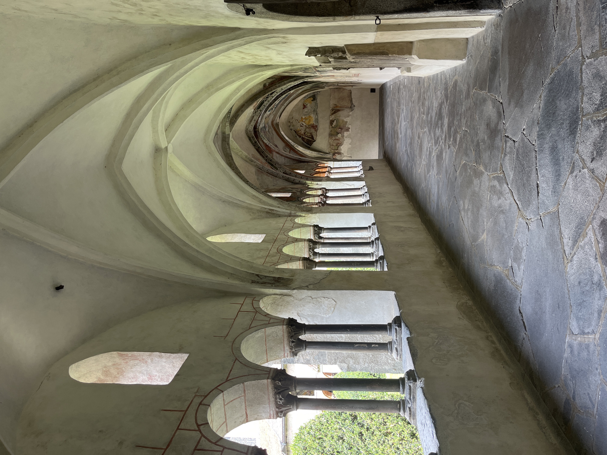 The image shows a cloister walkway with a stone floor and arched ceiling, featuring a row of columns separating the walkway from an outdoor garden. Frescoes adorn the walls of the cloister, depicting religious scenes. The architecture is historic, with a mix of stone and painted surfaces. The lighting suggests it is daytime, with natural light filtering in from the open side of the cloister. The overall impression is one of tranquility and historical significance, likely located within a monastery or cathedral complex. This scene is located in the cloister of Brixen Cathedral in Brixen (Bressanone), South Tyrol, Italy.