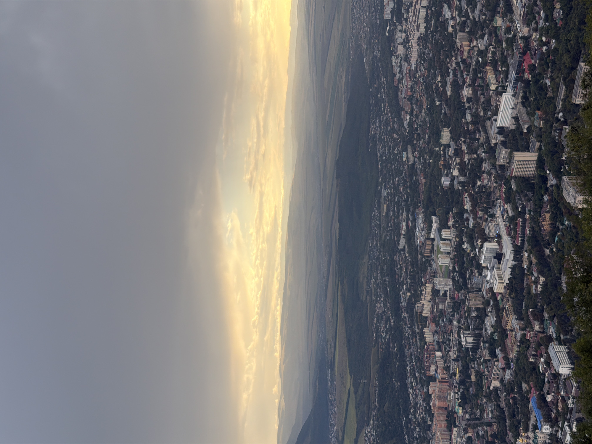 The image is an aerial view of the city of Goryachevodsky, Russia, taken during sunset. The foreground shows the dense urban landscape of Goryachevodsky, characterized by a mix of residential buildings, apartment blocks, and smaller structures, interspersed with abundant green trees. The buildings vary in architectural style and color, with some featuring balconies and modern designs, while others appear older. Roads and pathways are visible, indicating the city's layout.
In the middle ground, the urban area transitions into a more rural or semi-urban landscape with scattered vegetation and open fields. Further in the distance, rolling hills and mountains form the backdrop. The sky is a prominent feature, with dramatic clouds partially obscuring a bright, golden sunset. The warm hues of the setting sun cast a soft light across the horizon, creating a layered effect of light and shadow over the landscape. The overall impression is of a vast, sprawling city nestled within a natural environment, illuminated by the fading light of day. No people or specific actions are discernible from this high vantage point.