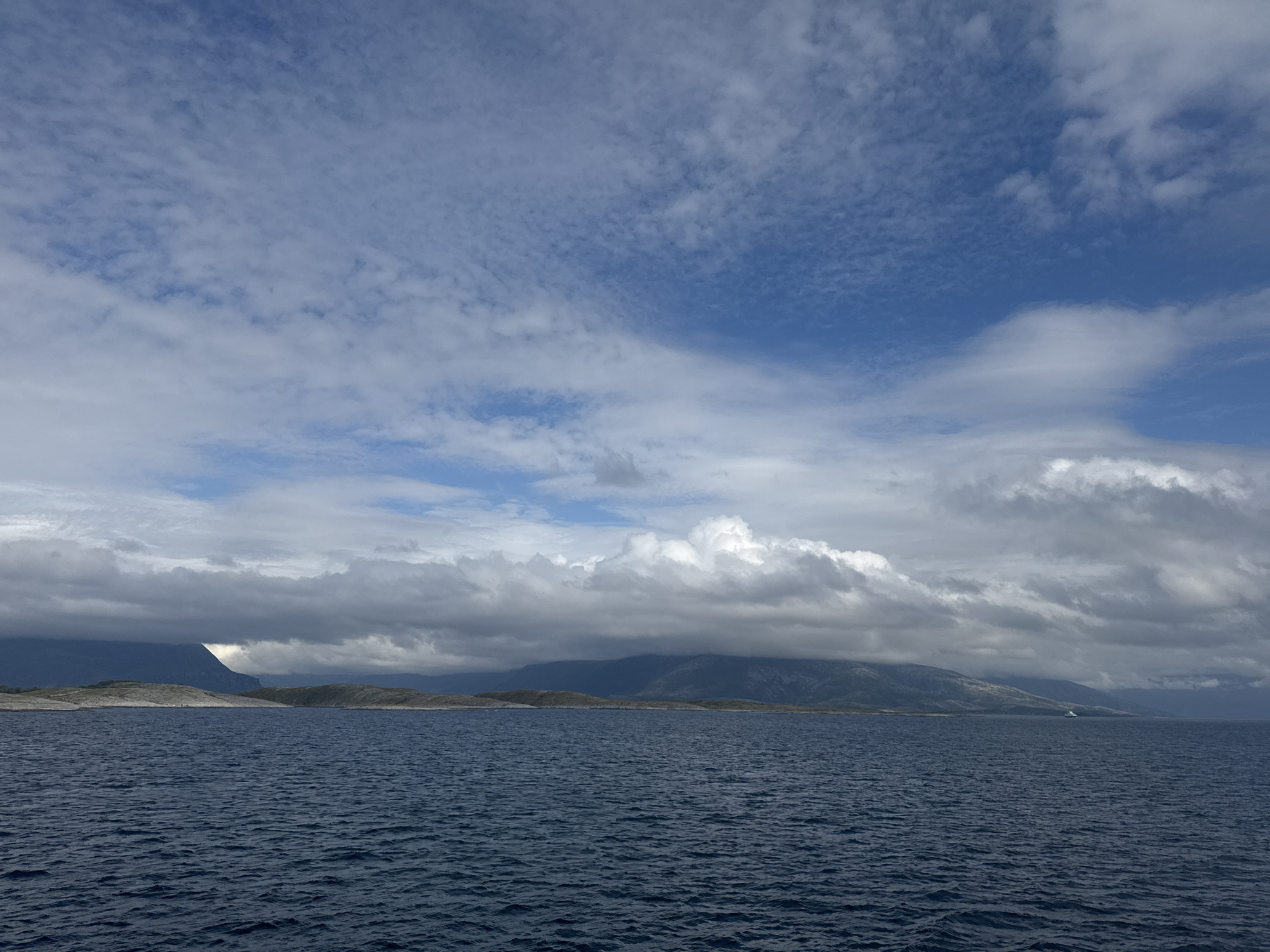 The image depicts a wide expanse of dark, choppy water stretching towards a distant, mountainous coastline under a dramatic sky. The sea occupies the bottom half of the frame, its surface rippled by waves. The horizon is marked by a low-lying, hilly landmass interspersed with rocky outcrops. Behind this, imposing mountains rise, partially obscured by a dense layer of dark, brooding clouds. These clouds create a sense of depth and shadow, with breaks allowing glimpses of a brighter sky and scattered lighter clouds above. The lighting suggests it could be late afternoon or early evening, with the sun's position casting subtle highlights on the distant peaks and illuminating some of the clouds from behind. There is a hint of a bright white patch on the left side of the mountain range, possibly a snow-covered peak or a glint of sunlight on rock. In the far distance, a small white vessel is visible on the water, a solitary speck against the vastness of the scene. The overall mood is serene yet powerful, evoking the rugged beauty of a coastal landscape.