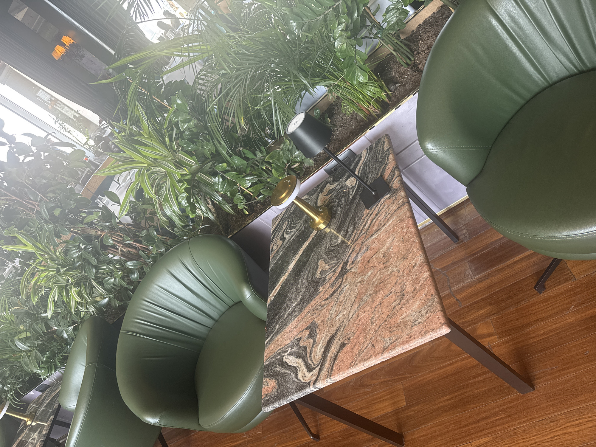 In the Sabail Raion, Azerbaijan, an indoor seating area is depicted. Three plush, dark green leather armchairs are arranged around a small, square table with a marble top. The table is supported by slim, dark metal legs and features two small lamps: one brass with a white shade, and another black with a bell-shaped shade. Behind the table and chairs, an abundance of lush green plants creates a natural backdrop, filling a long planter box. The flooring is polished dark wood. The overall impression is one of a modern and comfortable lounge space, possibly in a hotel lobby or upscale restaurant.