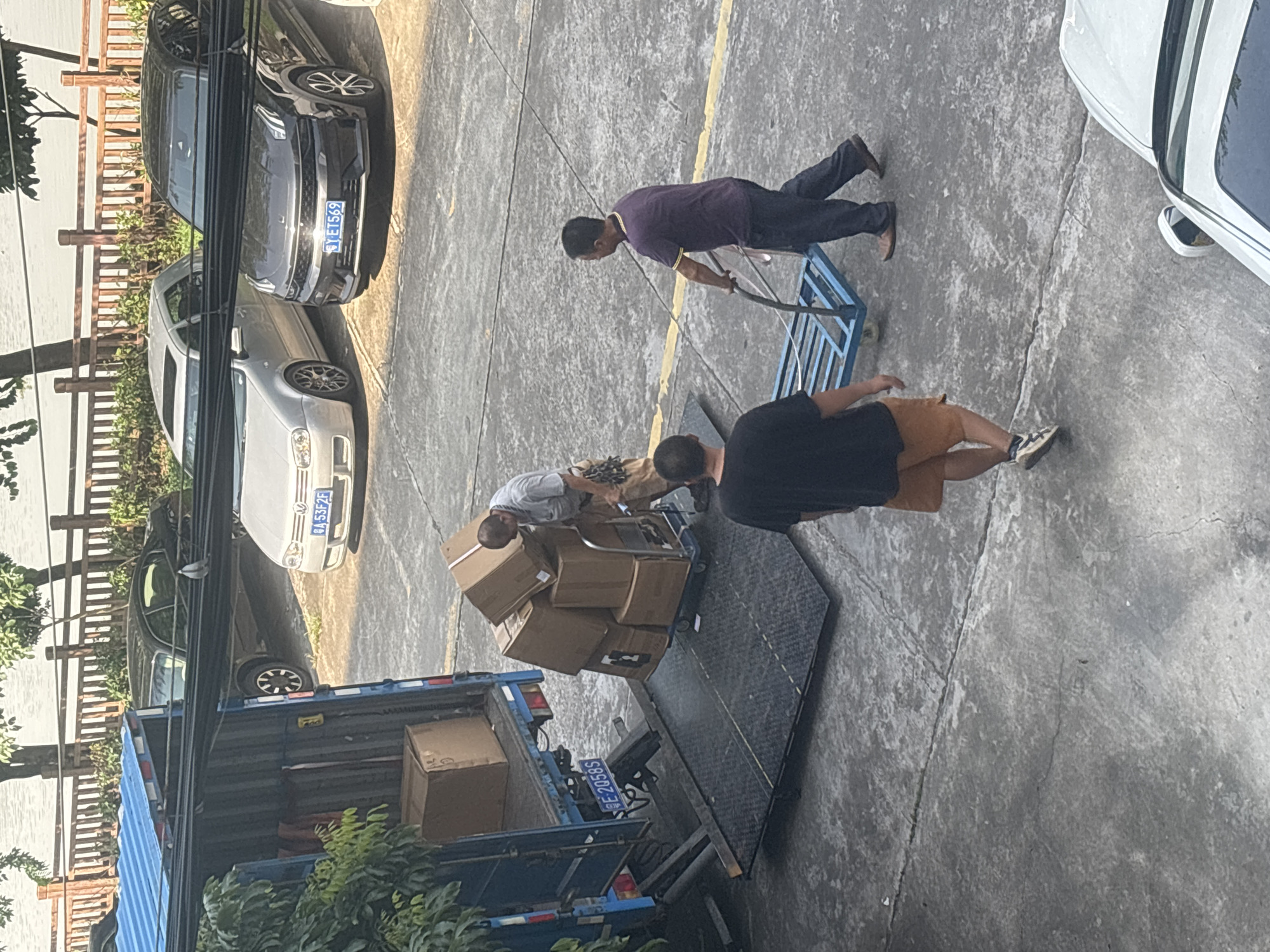 This is an eye-level, medium shot of a loading dock area in Nanhai District, China. The scene captures a delivery or move in progress, with several men engaged in the physical labor of transferring boxes from a blue truck. The truck is parked with its rear lift gate lowered, facilitating the movement of goods. A man in a grey shirt is actively stacking brown cardboard boxes onto a hand truck, which is positioned on the lift gate. Another man, wearing a dark purple polo shirt and dark pants, is pushing a blue metal cart. A third man, wearing a black t-shirt and orange shorts, walks away from the truck towards the foreground. The environment appears to be an urban outdoor space, possibly a commercial or residential complex, with a paved surface and a body of water visible in the background, separated by a wooden fence and some vegetation. Several cars, including a dark grey SUV and a silver hatchback, are parked in the vicinity. The lighting suggests it is daytime, with some shadows cast on the ground.