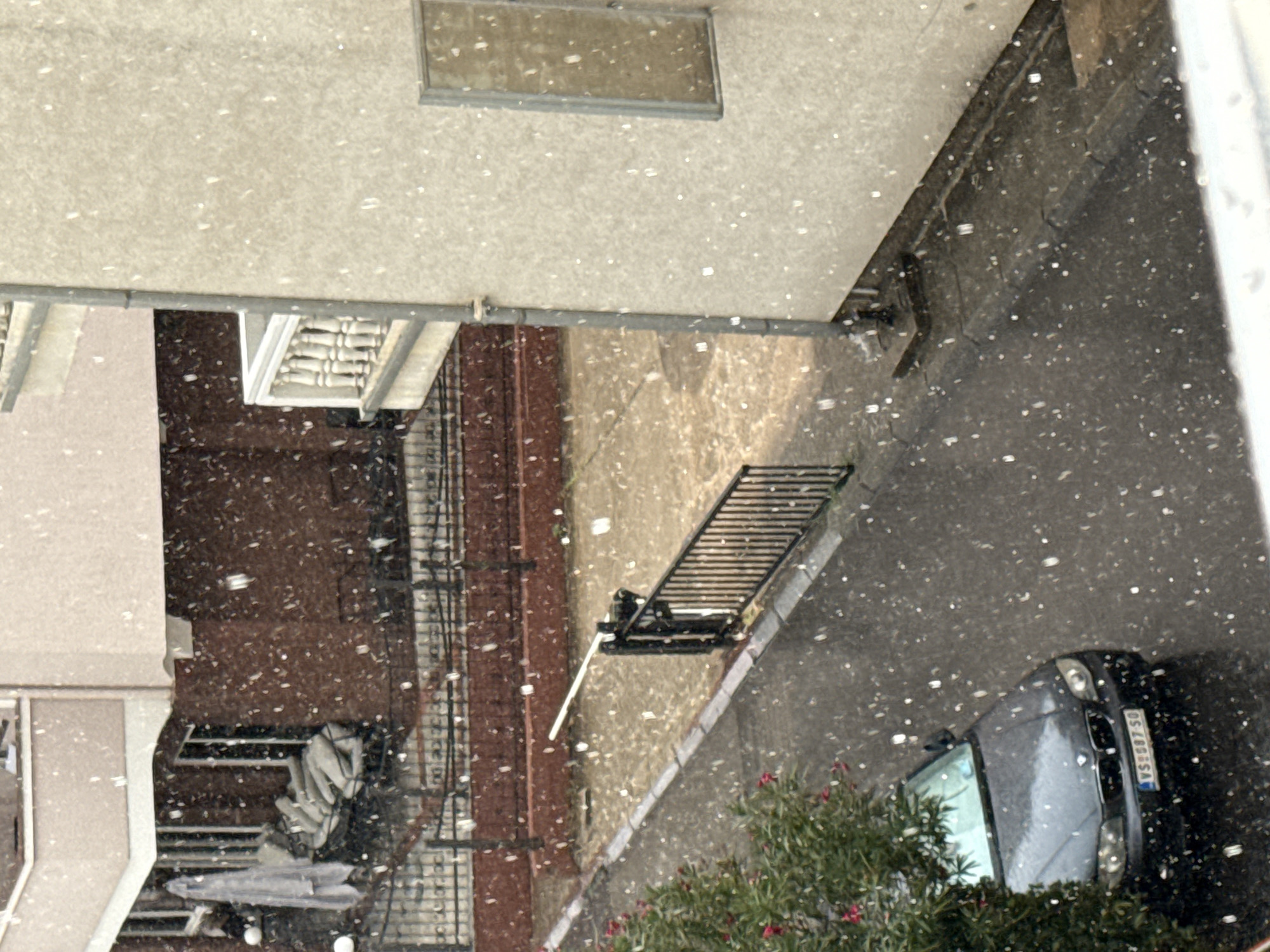 The image depicts a street scene in Budva, Montenegro, during a snowfall. A dark-colored sedan, identified by its license plate "VS 087 SO," is parked on the wet asphalt road. To the left, a green tree with red flowers is visible. In the background, a building with red-brown textured walls and a white balcony is partially obscured by the snowfall. A black metal gate with a white barrier arm is positioned across the road. The overall environment suggests an urban setting experiencing winter weather.