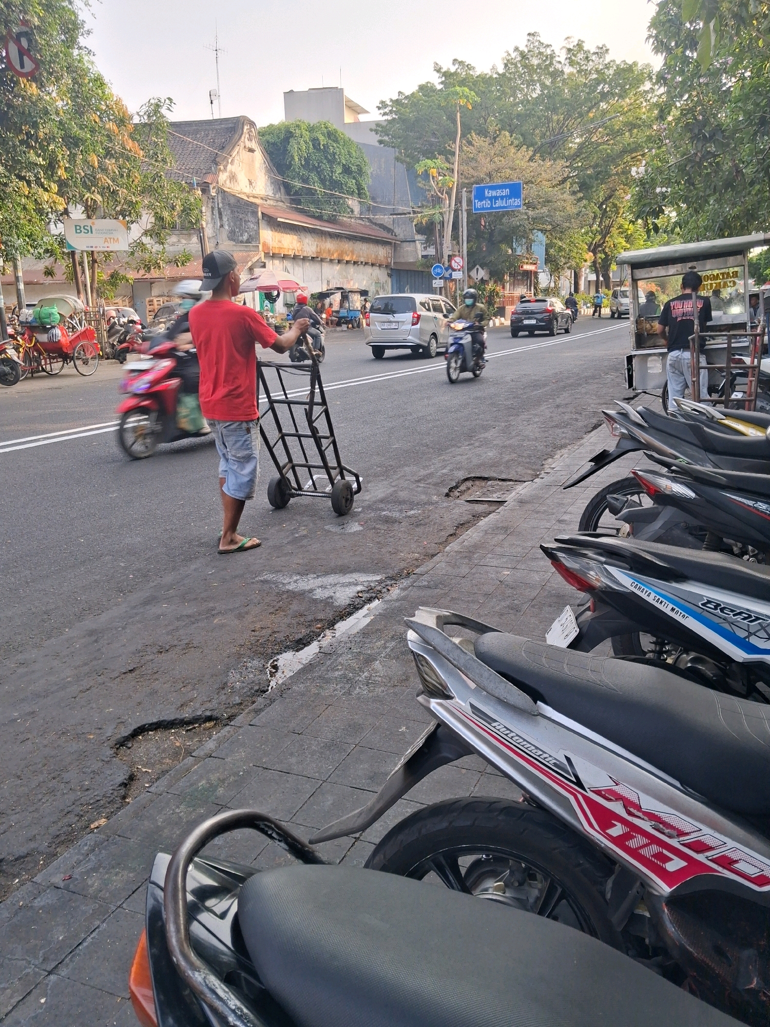 In Surakarta, Indonesia, on a street with several parked motorcycles and a food cart, a man in a red t-shirt and denim shorts pushes a handcart along the side of the road. A BSI ATM sign is visible on the left. Traffic, including a red motorcycle, a silver car, and other vehicles, moves along the street. A blue traffic sign reading "Kawasan Tertib Lalu Lintas" (Orderly Traffic Area) is positioned on the right side of the road. The scene is bathed in the warm light of what appears to be late afternoon or early morning.