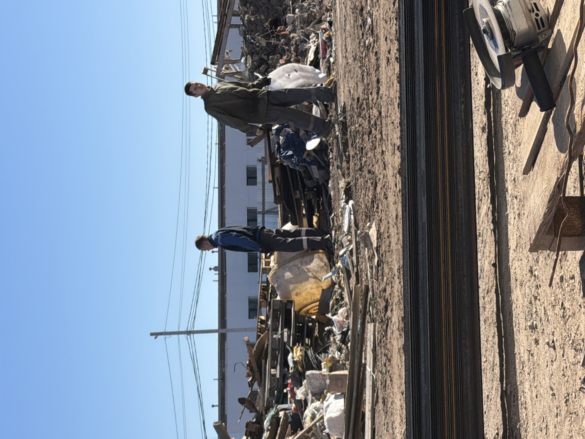 Two men stand in a cluttered, possibly industrial or construction site in what may be Chaikovsky, Russia. Debris and scrap materials litter the foreground and background. The men, dressed in work clothes, stand at opposite ends of a stack of metal beams. An angle grinder sits on the ground.
