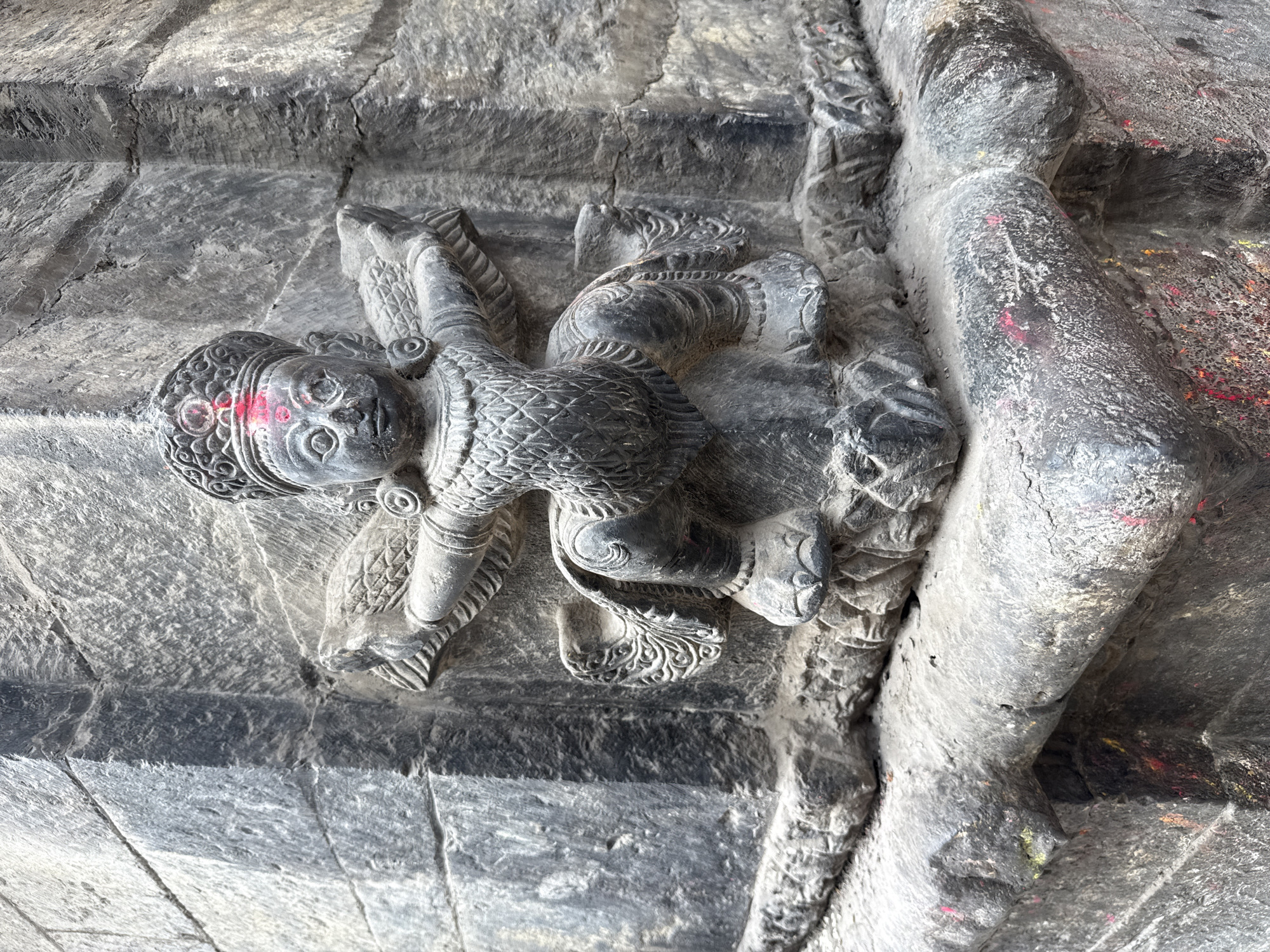 The image shows a stone carving of a figure on a wall, likely part of a temple structure. The figure has a round face, ornate headdress, and detailed body ornamentation. Red markings are visible on the face. The stone is grey and aged, exhibiting weathering and surface wear. The location is likely in Lalitpur, Nepal, known for its ancient temples and intricate stone carvings.