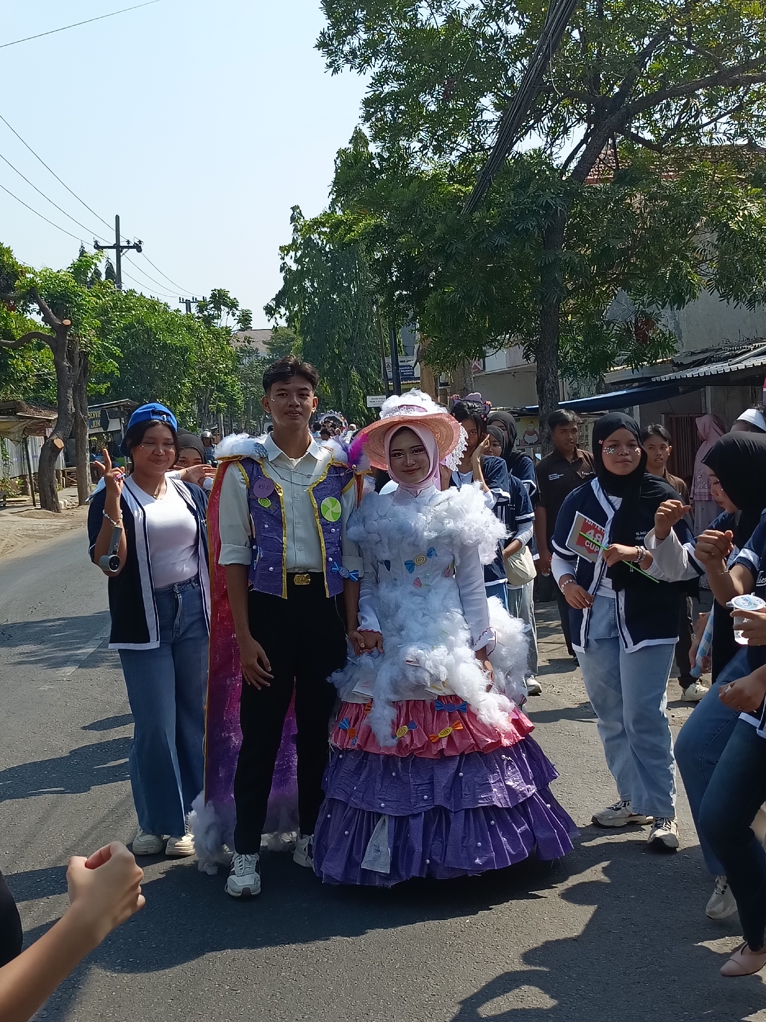 In the city of Kediri, Indonesia, a group of young people is participating in a parade on a paved road lined with trees and buildings. The central focus is a young man and woman dressed in elaborate costumes. The man wears a white collared shirt with a purple vest adorned with gold trim and circular decorations. He is also wearing black pants and a cape draped over his shoulders. The woman beside him is in a voluminous, multi-layered costume. Her dress features a white, fluffy top resembling clouds, a pink ruffled section, and a long purple skirt with a textured surface. She is also wearing a wide-brimmed pink hat and has glitter on her face.
Other individuals in the parade are visible in the background. To the left of the central pair, a young woman in a blue baseball cap and a white shirt with a dark jacket is making a peace sign with her fingers. Further to the right, other participants are seen in casual attire, some wearing dark blue shirts with white accents, and one individual wears a black hijab with glitter accents on her face. Some individuals are holding small flags or cups. The environment suggests a sunny day, with clear skies and dappled shadows on the road from the surrounding trees. The buildings in the background appear to be single or two-story structures, some with visible awnings or roofs. The scene captures a festive atmosphere, likely a school or community event.