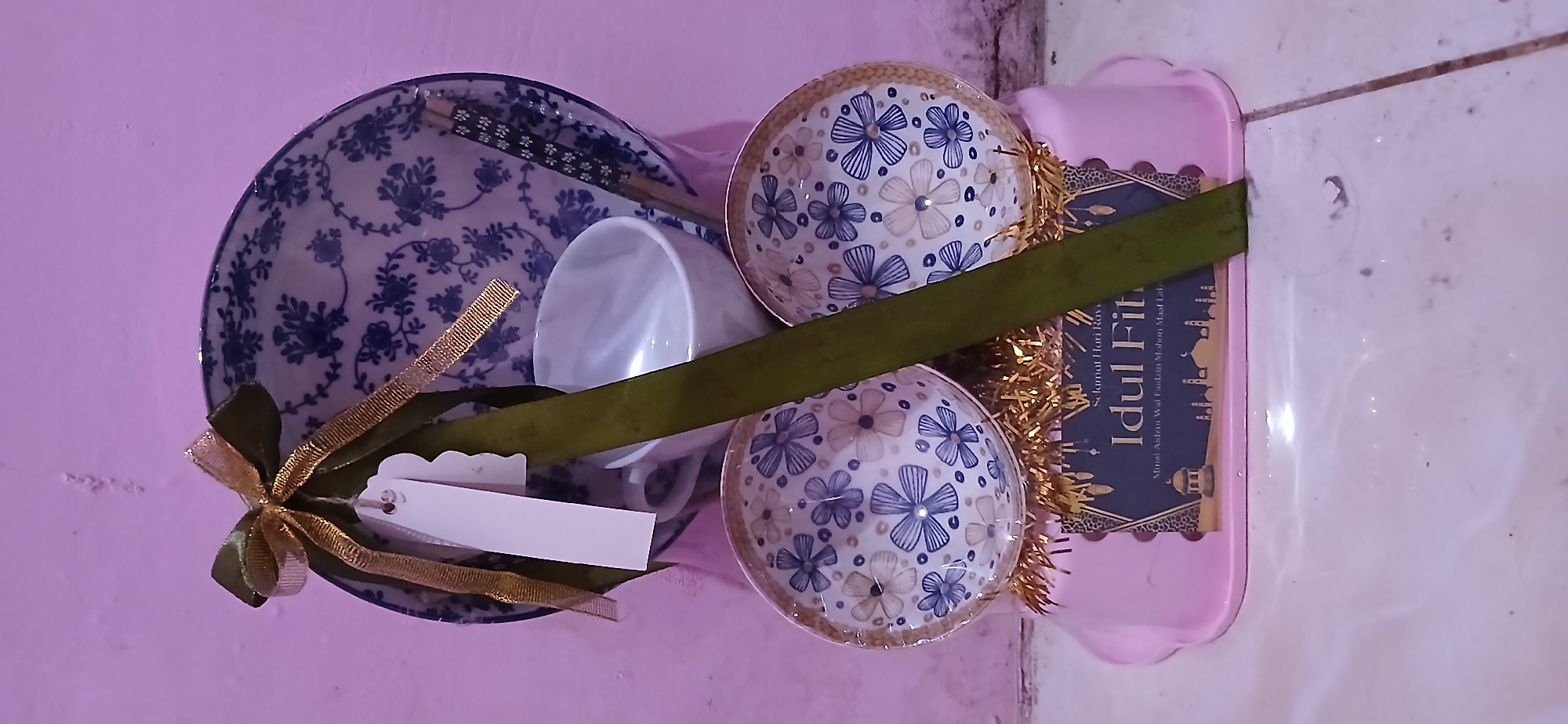 A gift basket is arranged in a vertical composition against a plain, pinkish wall. The basket, which appears to be made of pink plastic, contains several items. At the top is a large blue and white patterned plate, behind which a white teacup is visible. To the right of the teacup, a pair of chopsticks with a floral pattern is placed. In front of the large plate and teacup, two smaller blue and white patterned bowls are stacked. The bowls are placed on a bed of gold tinsel. A green ribbon is draped vertically in front of the objects, tied with a gold ribbon and a white tag at the top. Below the bowls and tinsel, a dark blue decorative card with gold accents displays text in Indonesian, including "Selamat Hari Raya" and "Idul Fitri." The overall presentation suggests a gift intended for the Eid al-Fitr celebration. The lighting is dim, and the background wall shows some texture and imperfections. The floor in the foreground appears to be tiled. The image does not contain any people or provide any specific visual cues about the location in Samarinda, Indonesia, beyond the text on the card, which indicates an Indonesian context.