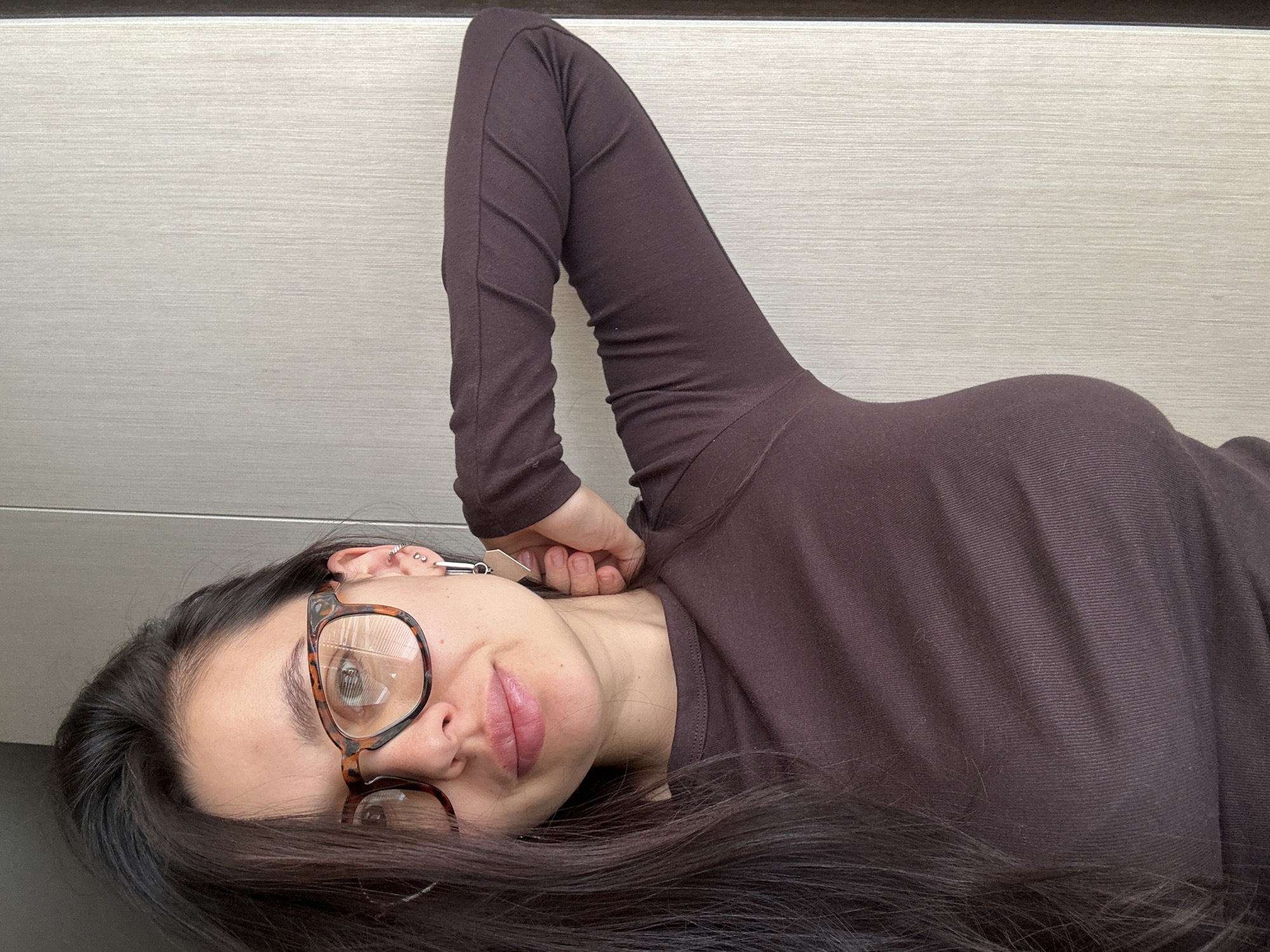 A woman is depicted lying on her side in an indoor setting, looking towards the camera. She has long, dark hair spread around her head and is wearing glasses with tortoiseshell frames. Her left eye is clearly visible through the lens, while her right eye is partially obscured. Her lips are slightly parted. Multiple small, metallic piercings adorn her left ear. She is dressed in a dark brown, long-sleeved top, which appears to have a ribbed or textured fabric. Her right arm is bent, with her hand resting near her head or neck, partially covered by her hair and clothing. The background is a light-colored, possibly wood or laminate, surface with a prominent horizontal grain. A dark, thin horizontal line is visible at the top edge of the frame, suggesting a border or transition in the surface. The woman's expression is calm and neutral, with a hint of a soft smile. There is no visible text in the image. The provided location context for this image is Chaikovsky, Russia.