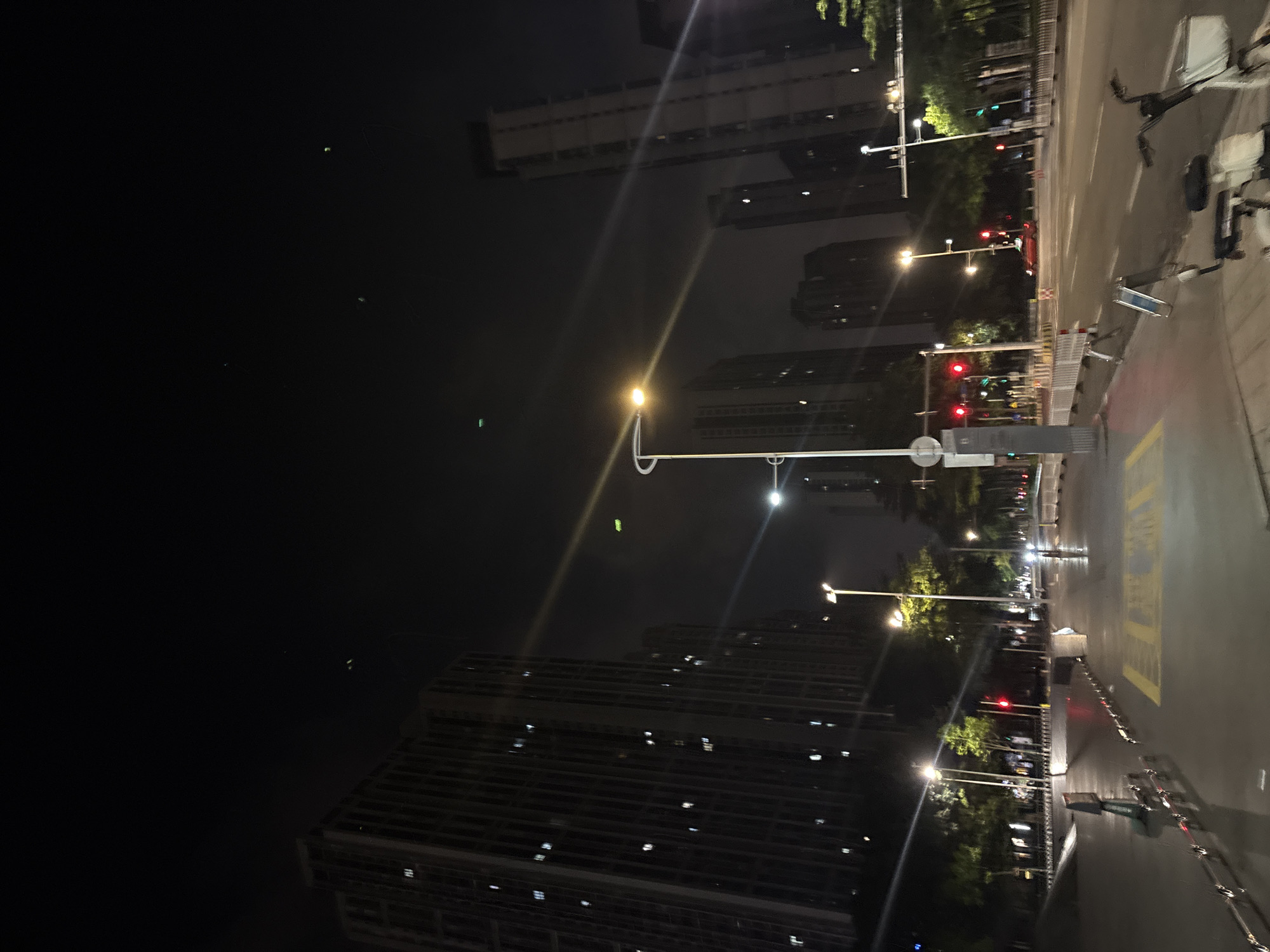The image depicts a nighttime scene in Haizhu District, China. Tall residential buildings with illuminated windows dominate the left side of the frame, contrasting with the dark sky above. Streetlights cast pools of yellow light on the wet pavement of a sidewalk and road. Traffic lights are visible, some showing red, indicating a traffic stop. Several trees line the street, their leaves illuminated by the artificial lights. A bicycle is parked on the sidewalk in the foreground. The overall impression is of a quiet, urban environment at night.