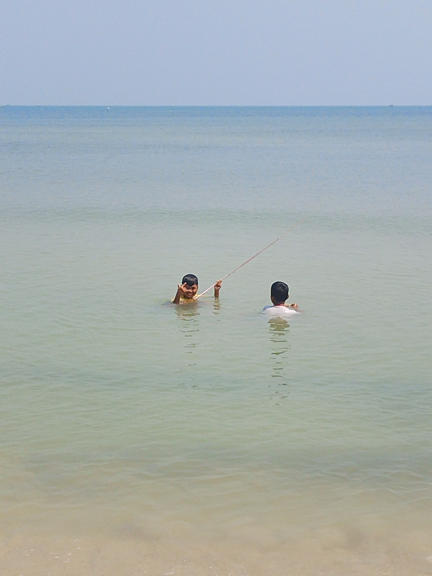 Two children are in the shallow ocean water near the shore. The child on the left, facing the camera, is wearing a yellow shirt and is making a peace sign with their fingers. This child is holding a thin, red string that extends to the right. The child on the right, facing away from the camera, is wearing a white shirt. The water is a pale green and calm, with the sandy shore visible at the bottom of the frame. The sky is a clear, pale blue. The image was taken on a sunny day. The location is Tegalsambi, Indonesia.